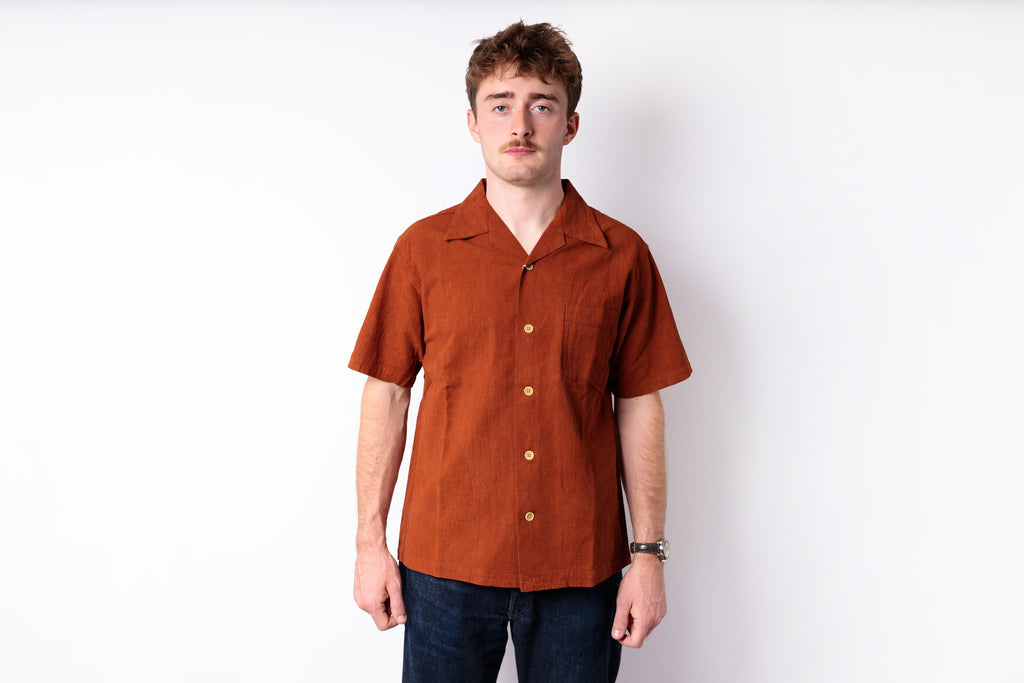 A man wearing a rust-orange short-sleeve linen camp collar shirt with gold buttons, paired with dark blue jeans, standing against a plain white studio background.
