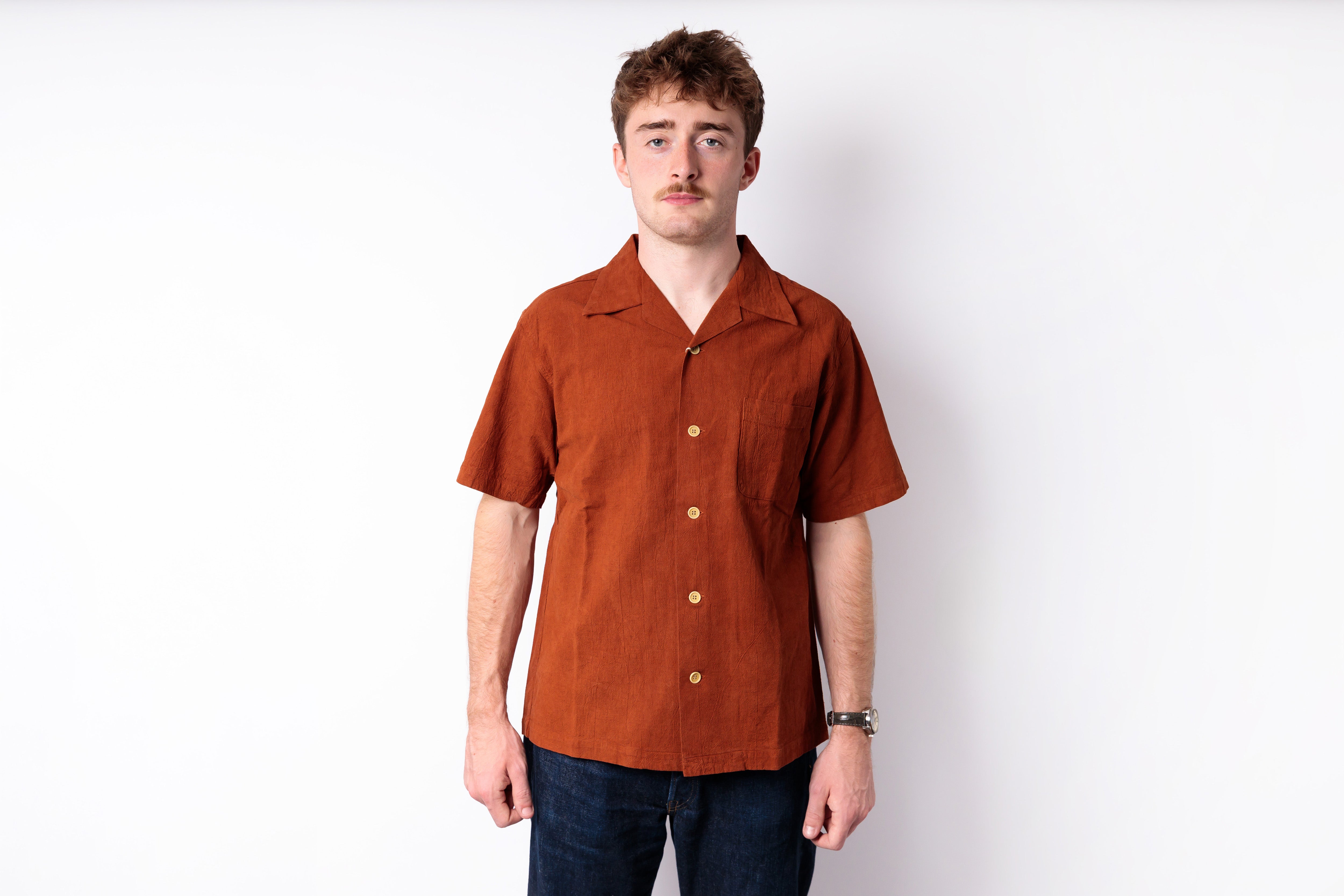A man wearing a rust-orange short-sleeve linen camp collar shirt with gold buttons, paired with dark blue jeans, standing against a plain white studio background.
