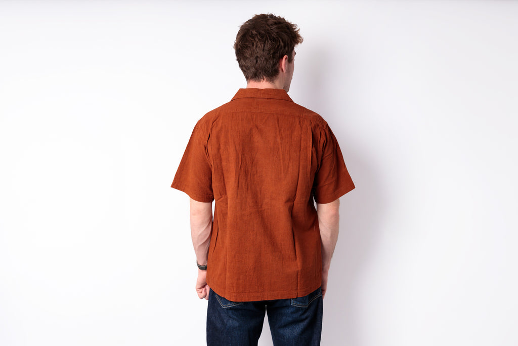 A man viewed from behind wearing a rust-orange short-sleeve linen shirt and dark blue jeans, standing against a plain white wall.