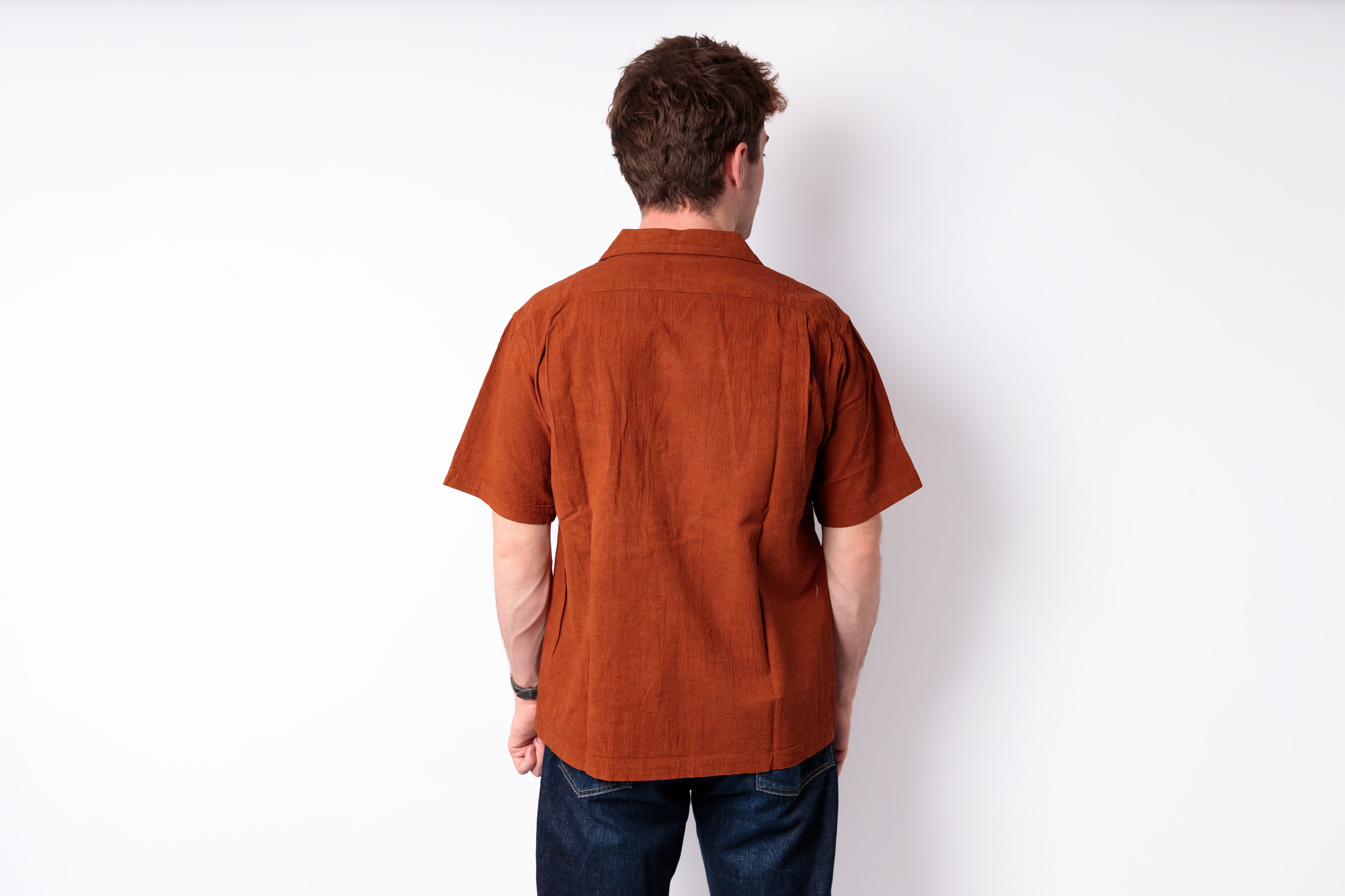 A man viewed from behind wearing a rust-orange short-sleeve linen shirt and dark blue jeans, standing against a plain white wall.