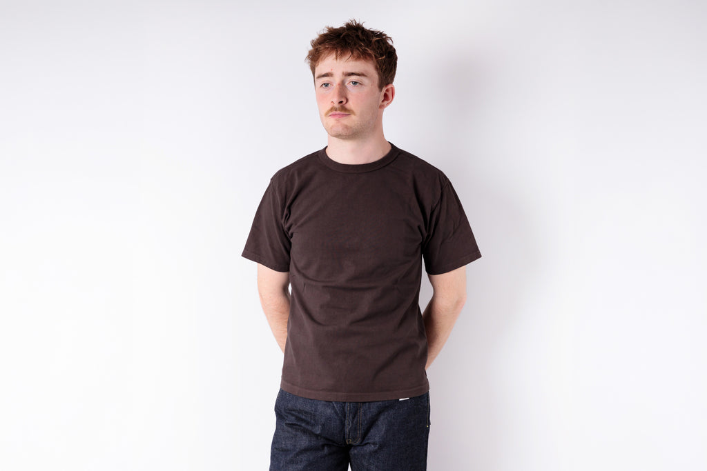 Man wearing a brown t-shirt and dark jeans against a white background