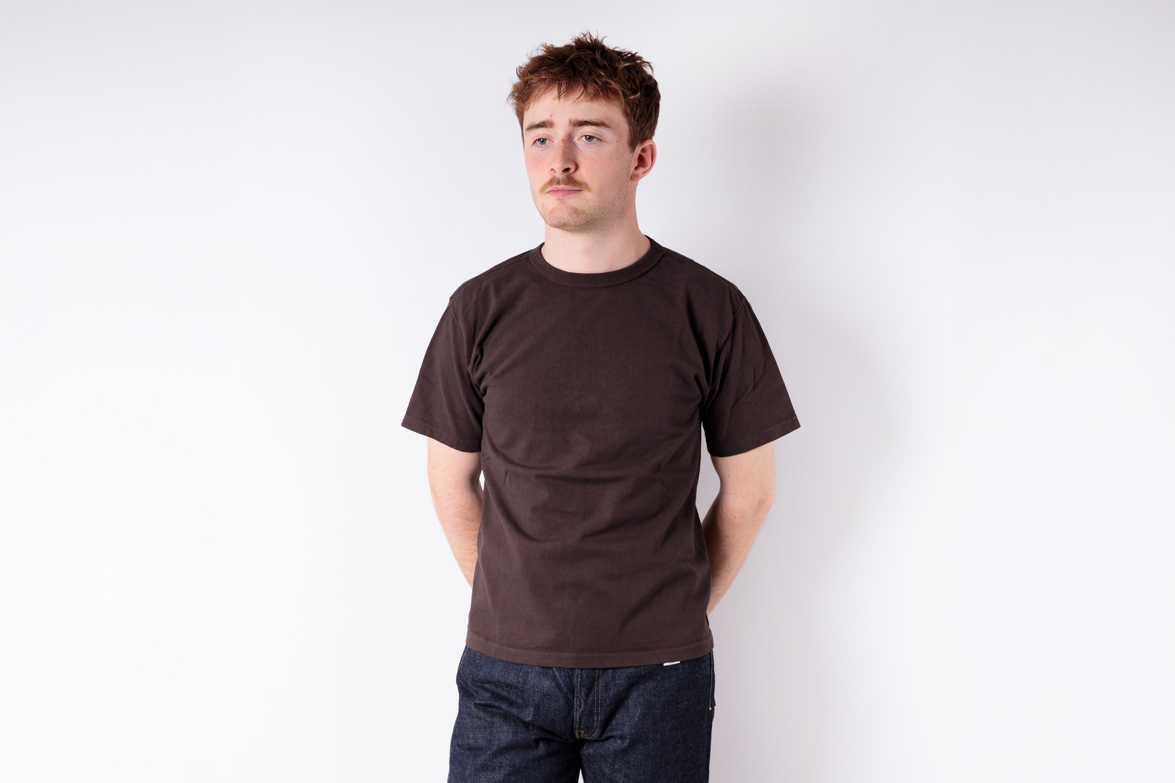 Man wearing a brown t-shirt and dark jeans against a white background