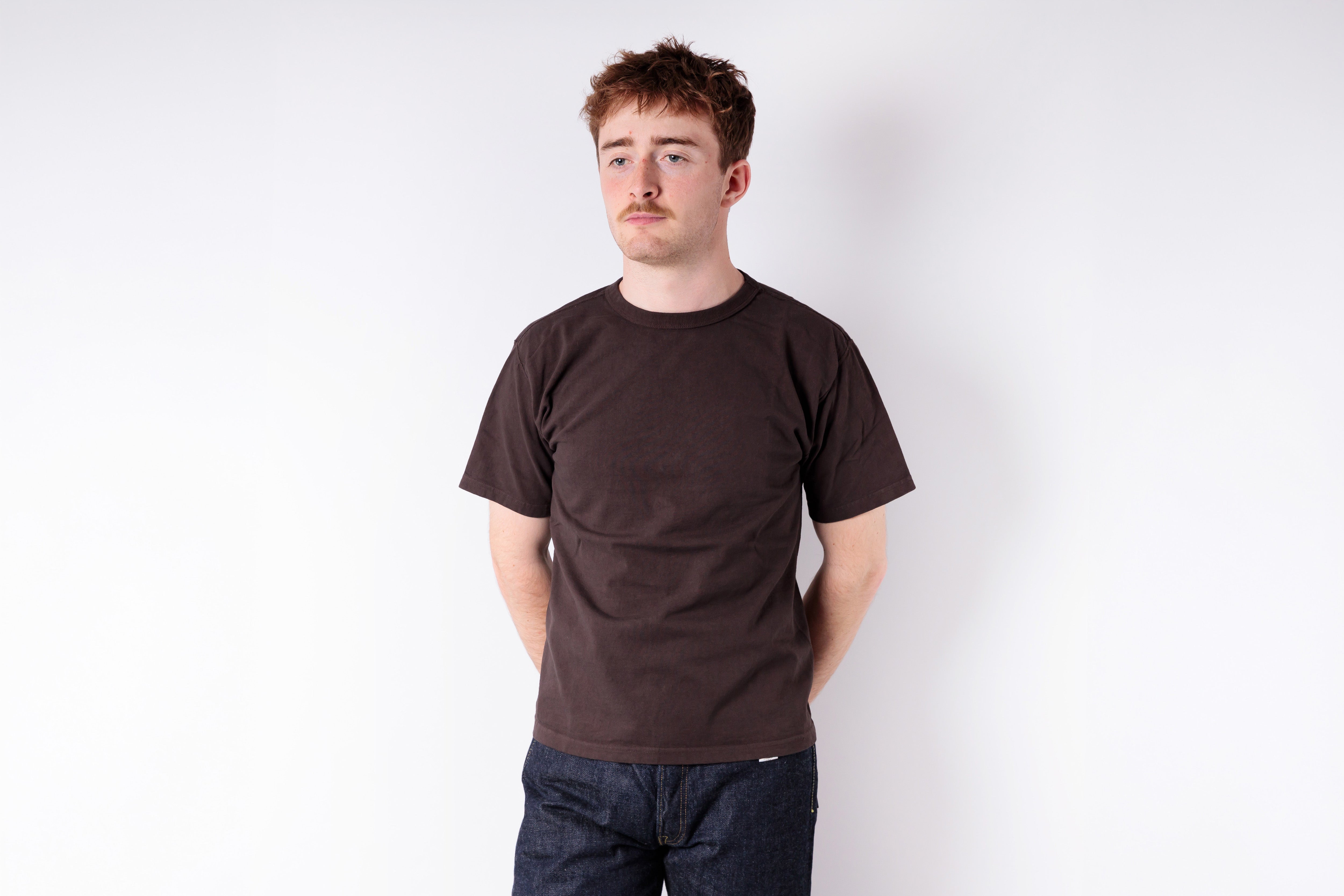 Man wearing a brown t-shirt and dark jeans against a white background