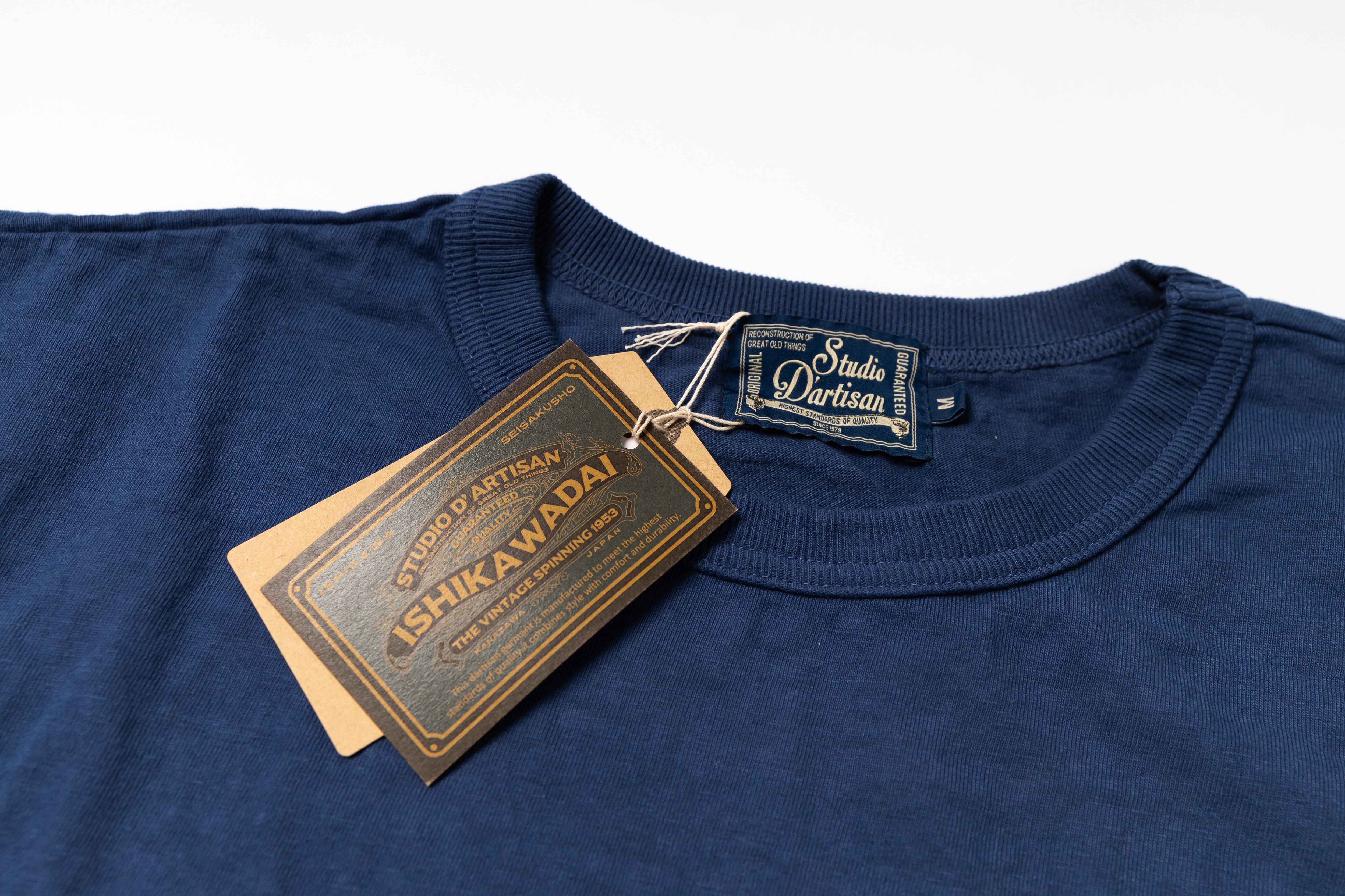 A close-up of a navy blue crewneck t-shirt with a 'Studio Partisan' neck label and a hanging 'Shikawadai' paper tag, set against a plain white background.