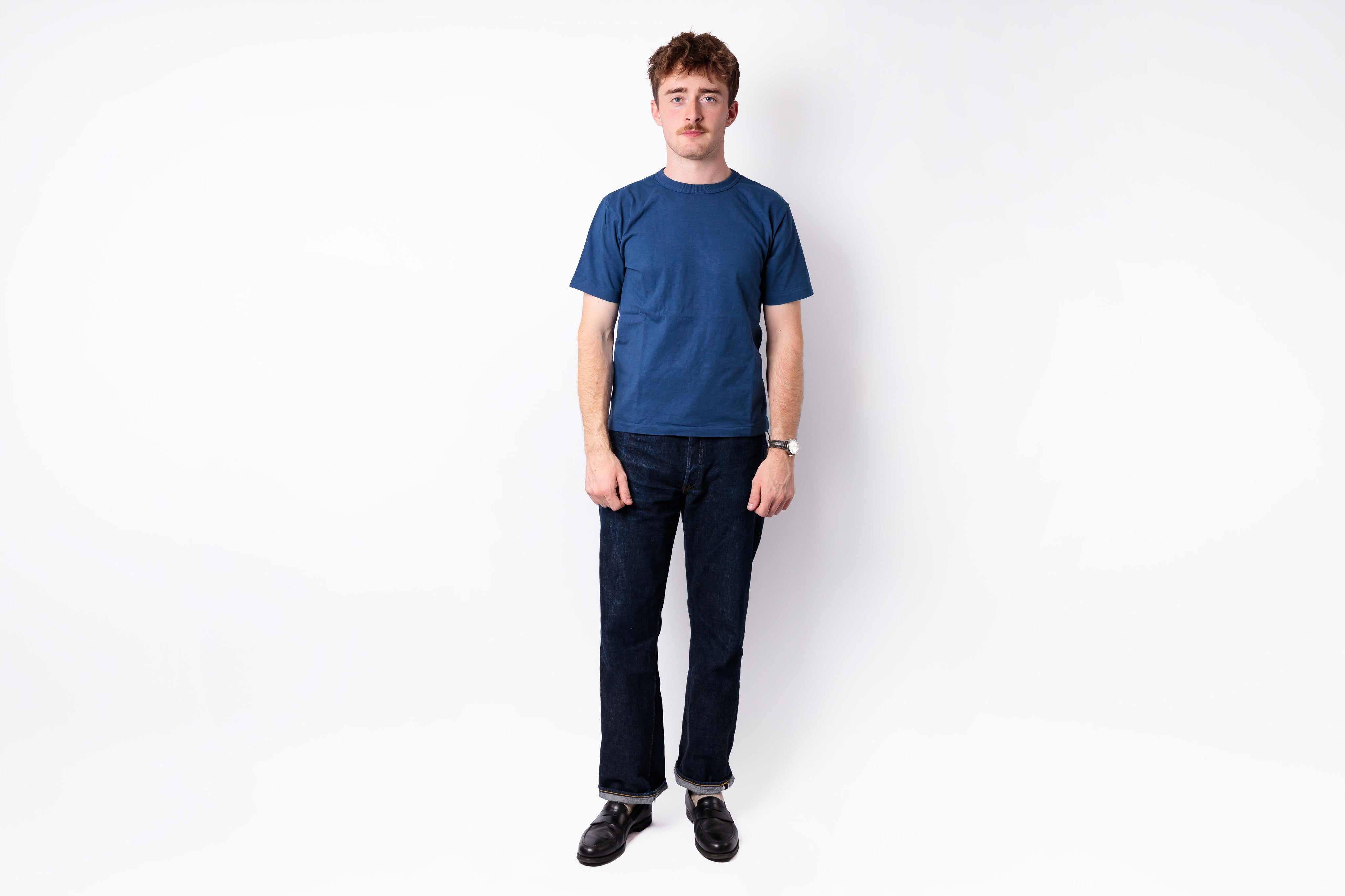 A man wearing a solid blue short-sleeve crewneck t-shirt, dark blue jeans, and black shoes, standing against a plain white studio background.