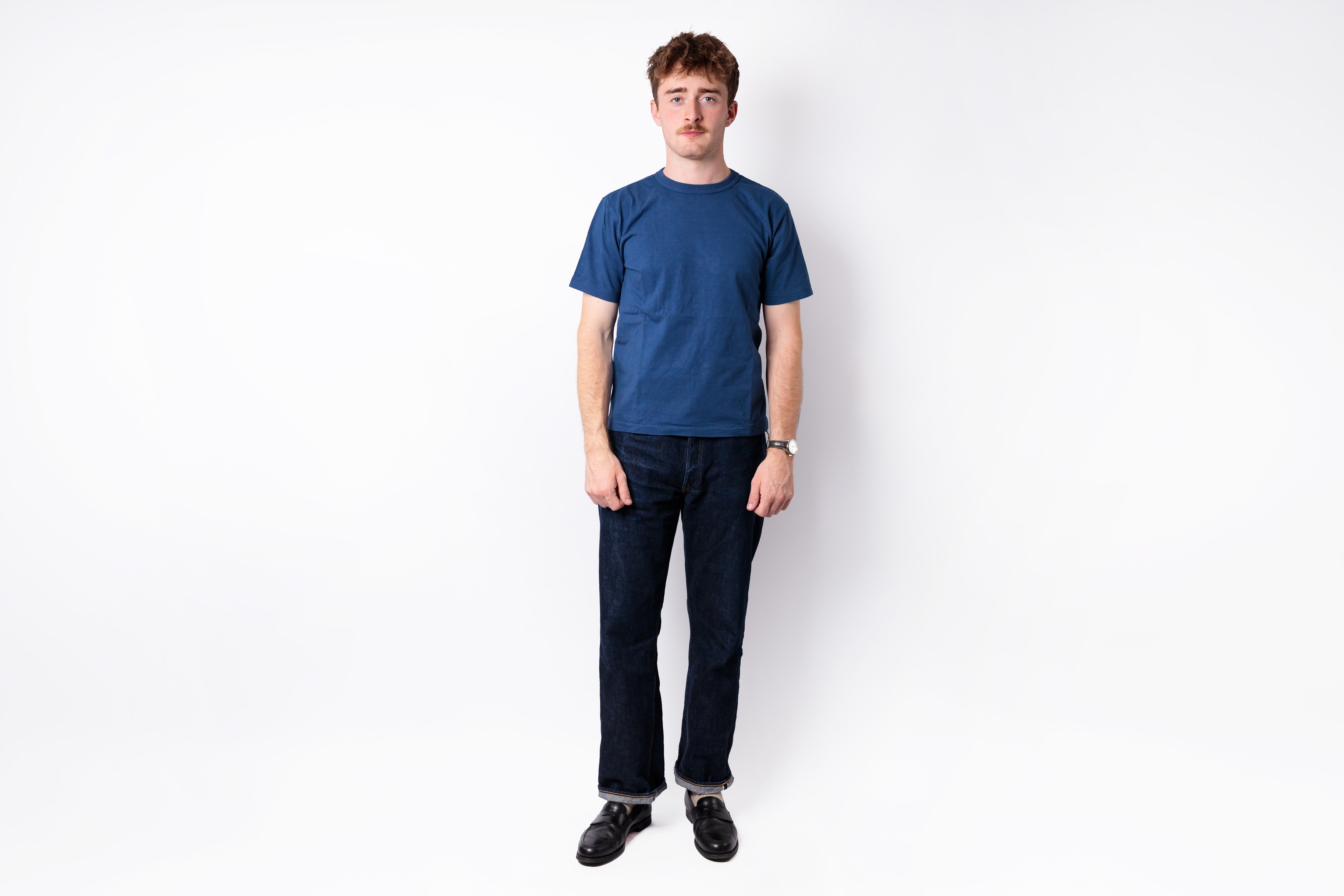 A man wearing a solid blue short-sleeve crewneck t-shirt, dark blue jeans, and black shoes, standing against a plain white studio background.