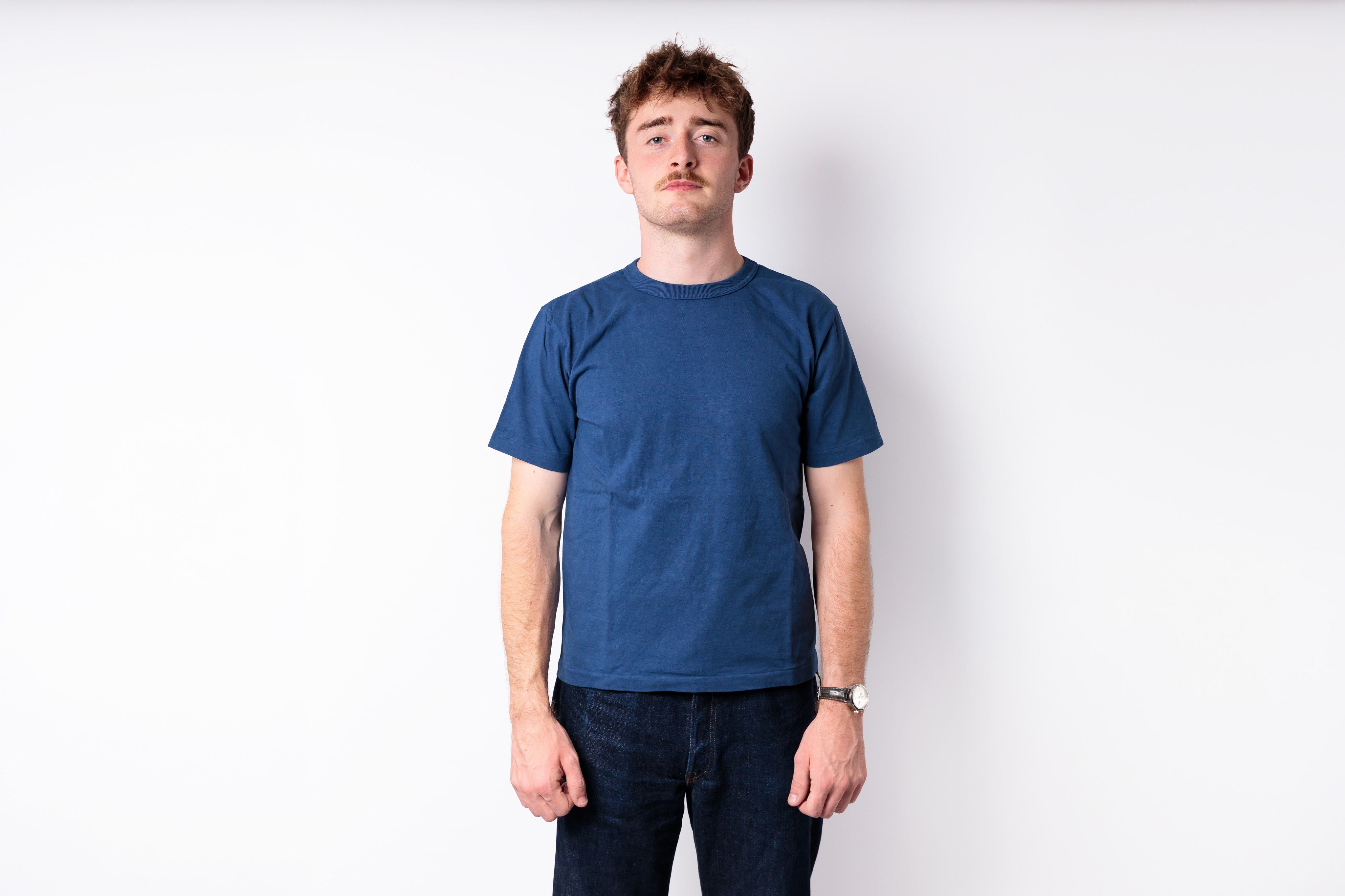 A man wearing a solid blue crewneck t-shirt and dark jeans, standing against a plain white studio background.