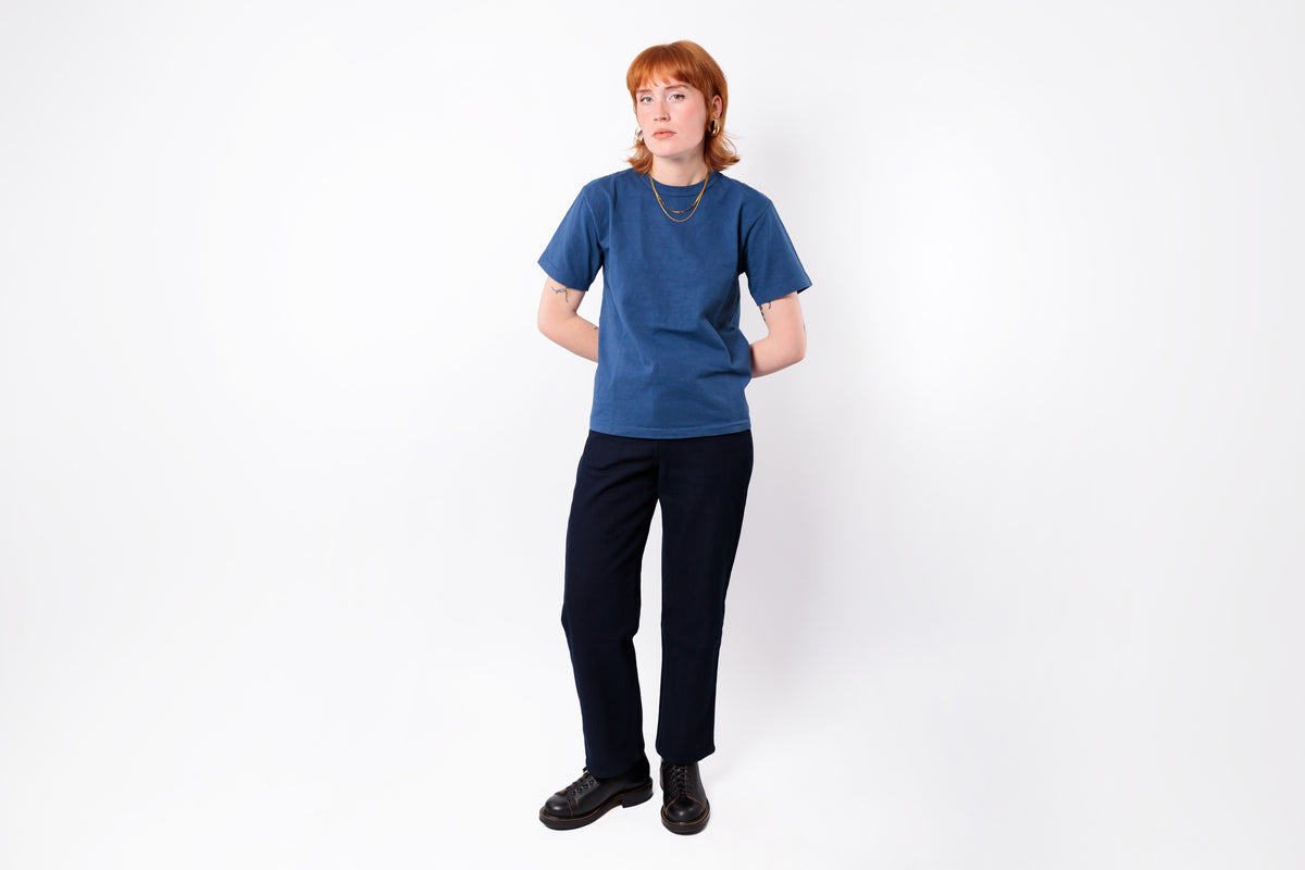 A person with red hair stands against a plain white studio background wearing a blue short-sleeve crewneck t-shirt, dark navy trousers, and black shoes.