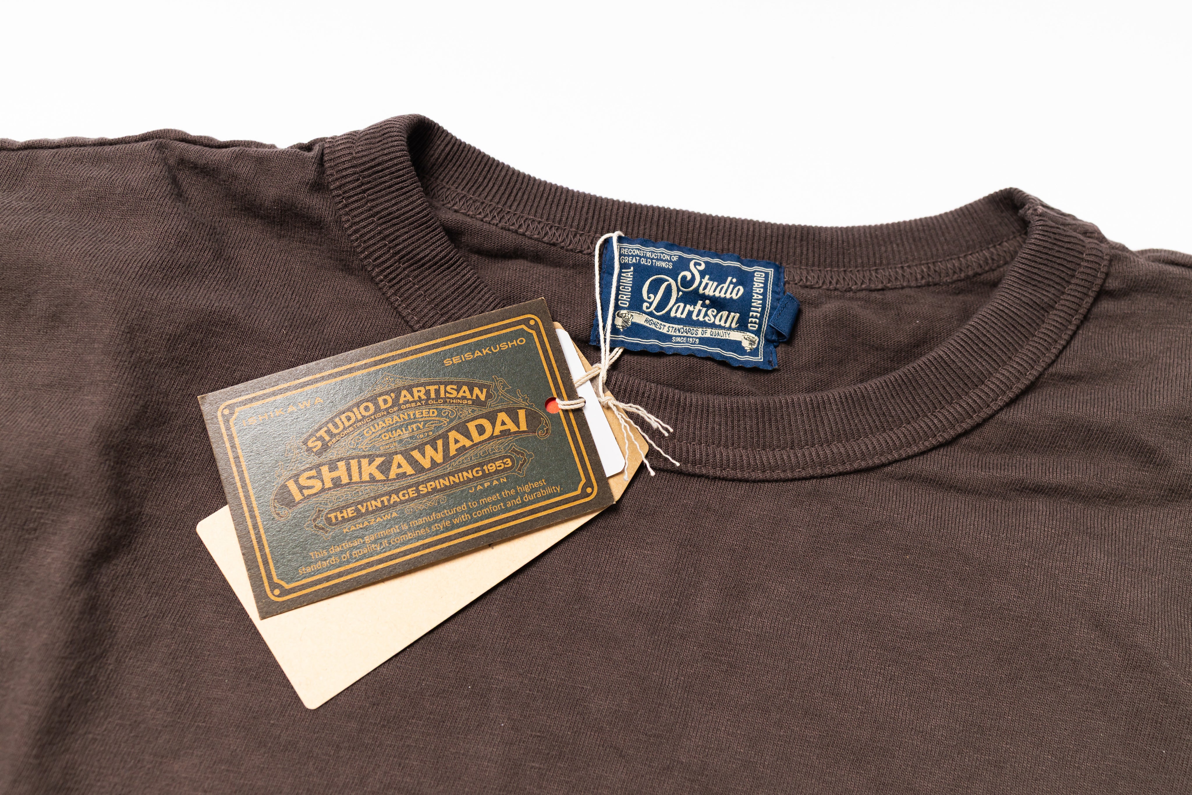 A close-up of a dark brown ribbed t-shirt with its original paper hangtag and a blue neck label. 