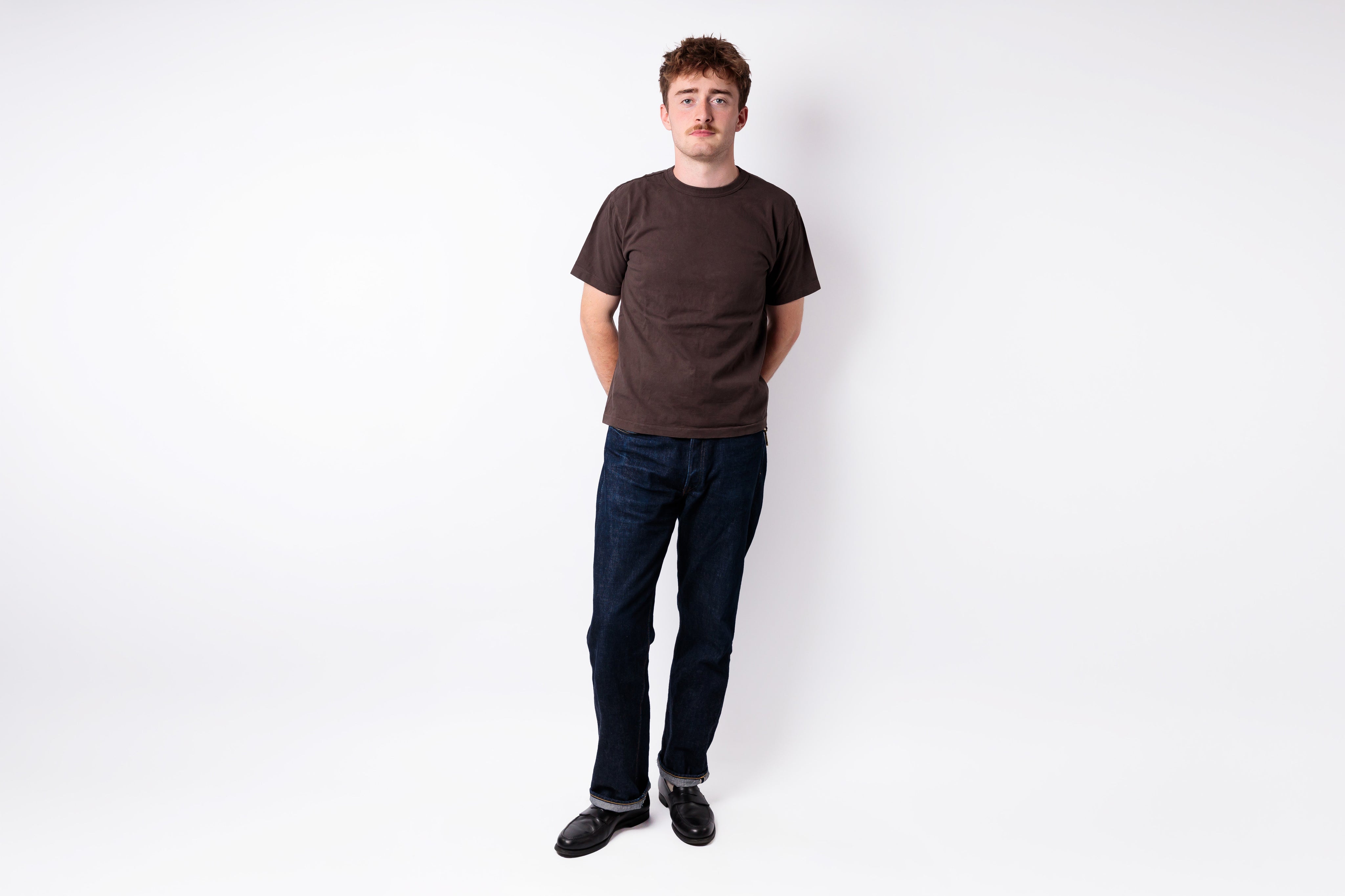 A man standing against a plain white studio background, wearing a dark brown short-sleeve crewneck t-shirt, dark blue jeans, and black shoes.