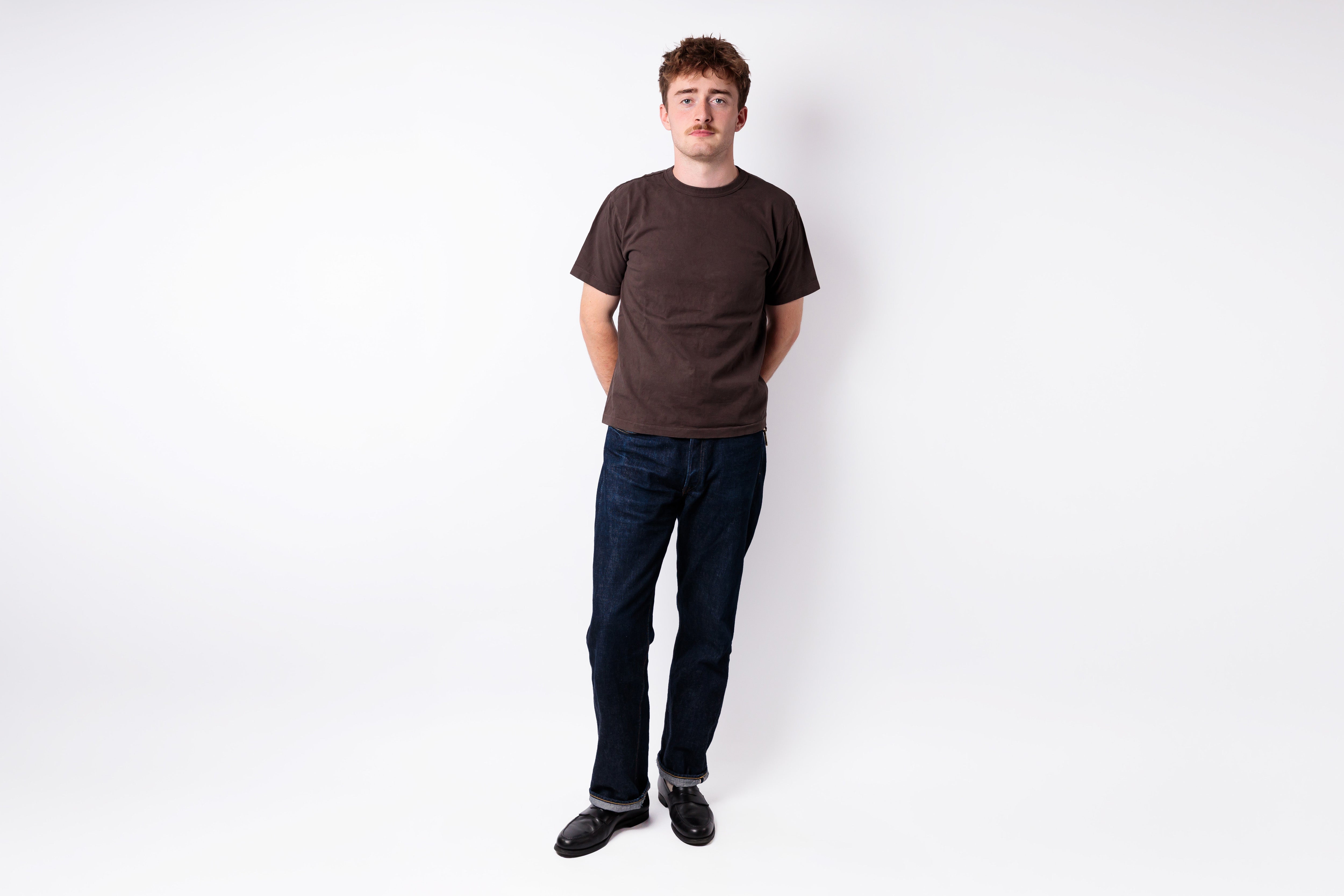 A man standing against a plain white studio background, wearing a dark brown short-sleeve crewneck t-shirt, dark blue jeans, and black shoes.