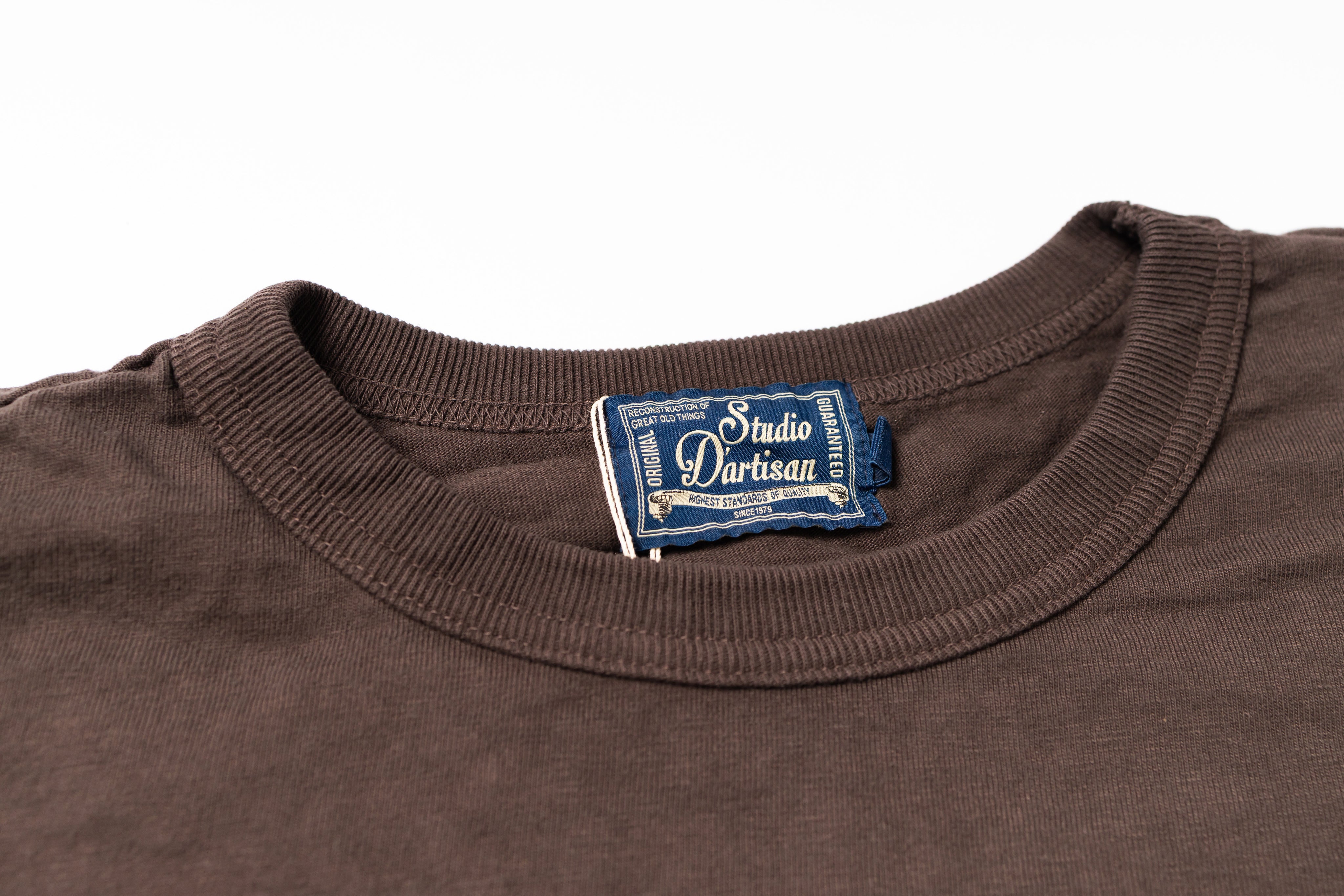 Close-up of a dark brown crewneck t-shirt showing the inner neck label which reads 'Studio D'Artisan'.