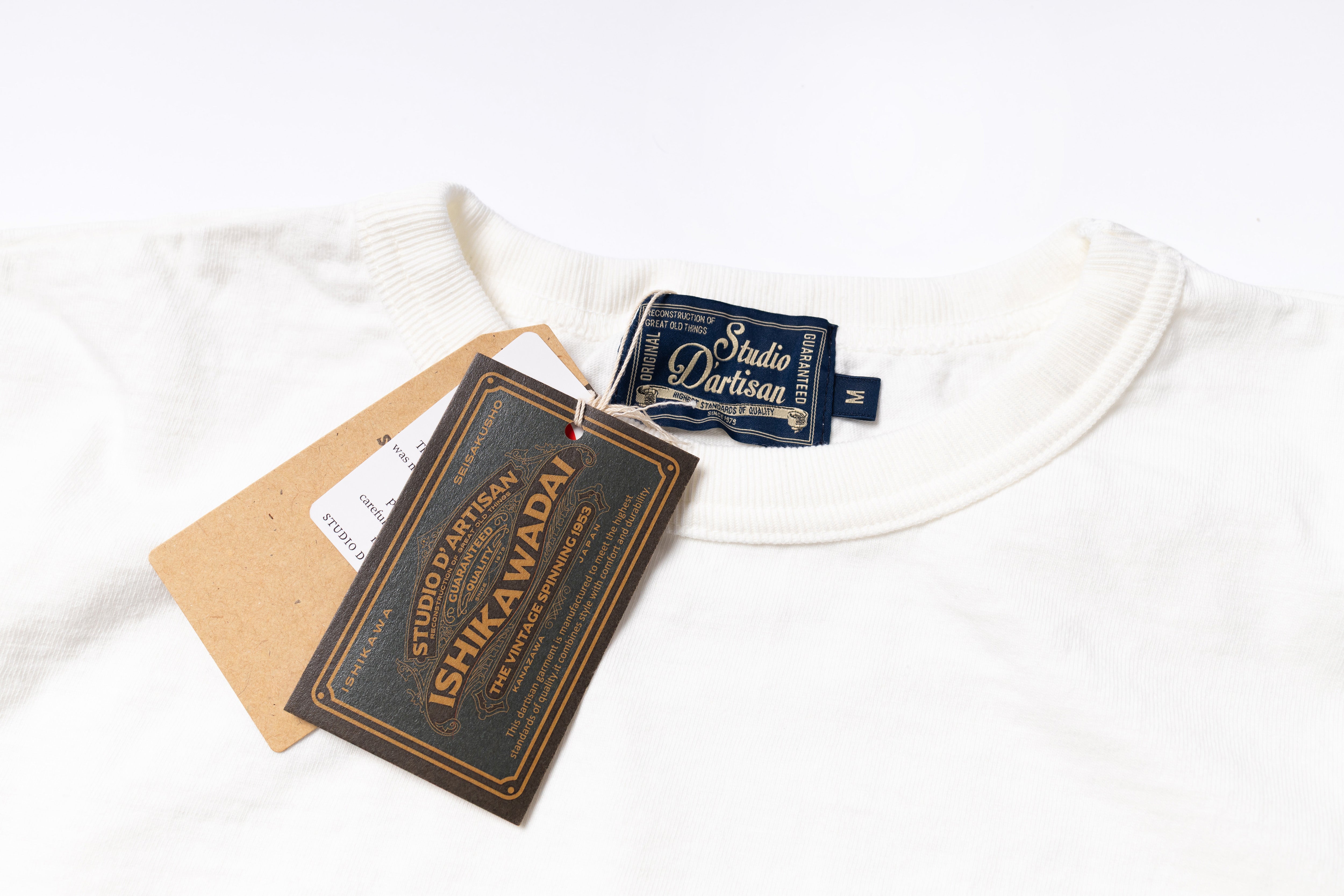 A close-up of a white crewneck sweatshirt laid flat on a white background, showing a dark blue neck label reading Studio Dartisan and kraft paper hang tags, including a prominent tag that reads Ishikawadai.