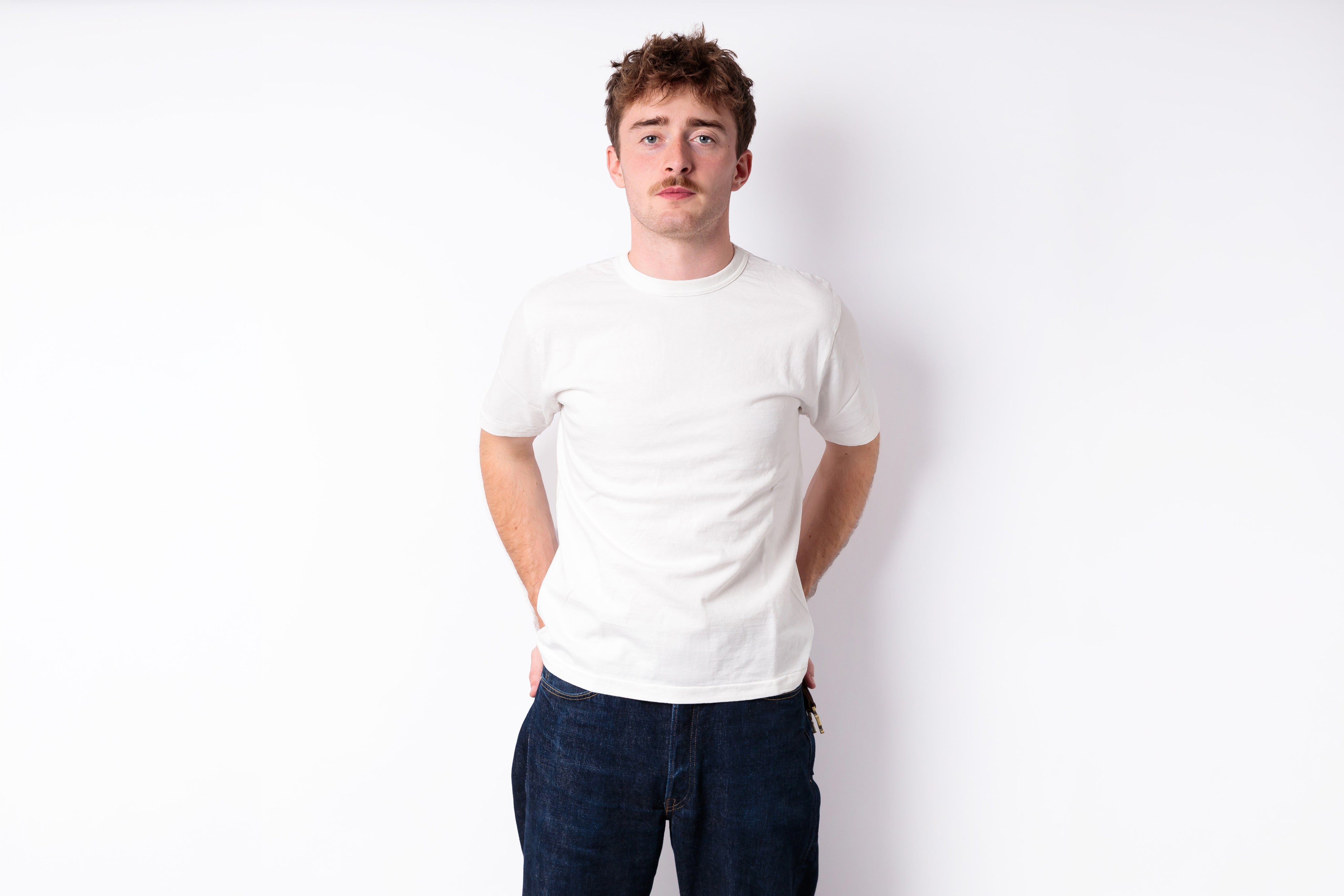 A young man with short brown hair and a mustache wearing a plain white crewneck t-shirt and dark blue jeans, standing against a solid white background.