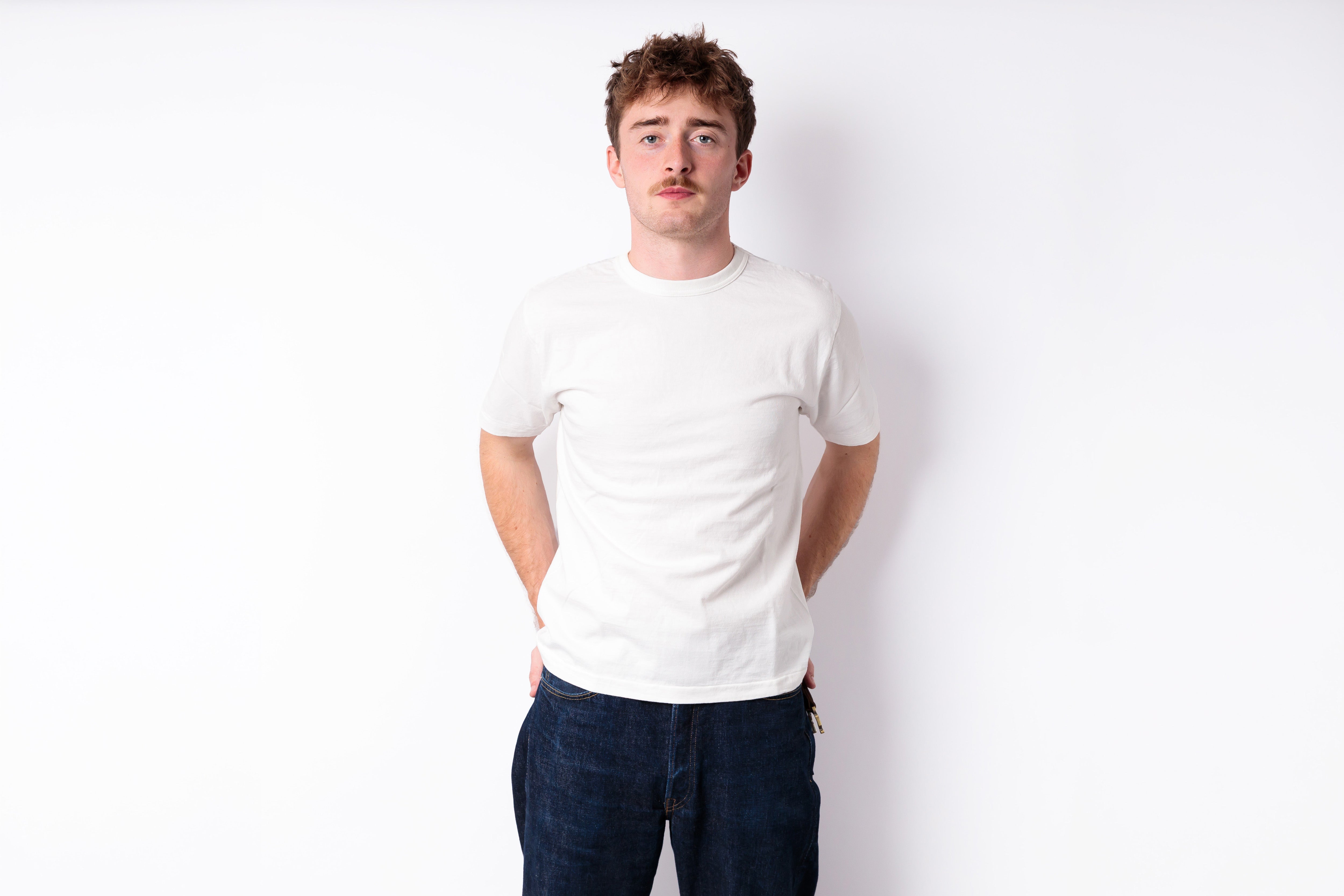 A young man with short brown hair and a mustache wearing a plain white crewneck t-shirt and dark blue jeans, standing against a solid white background.