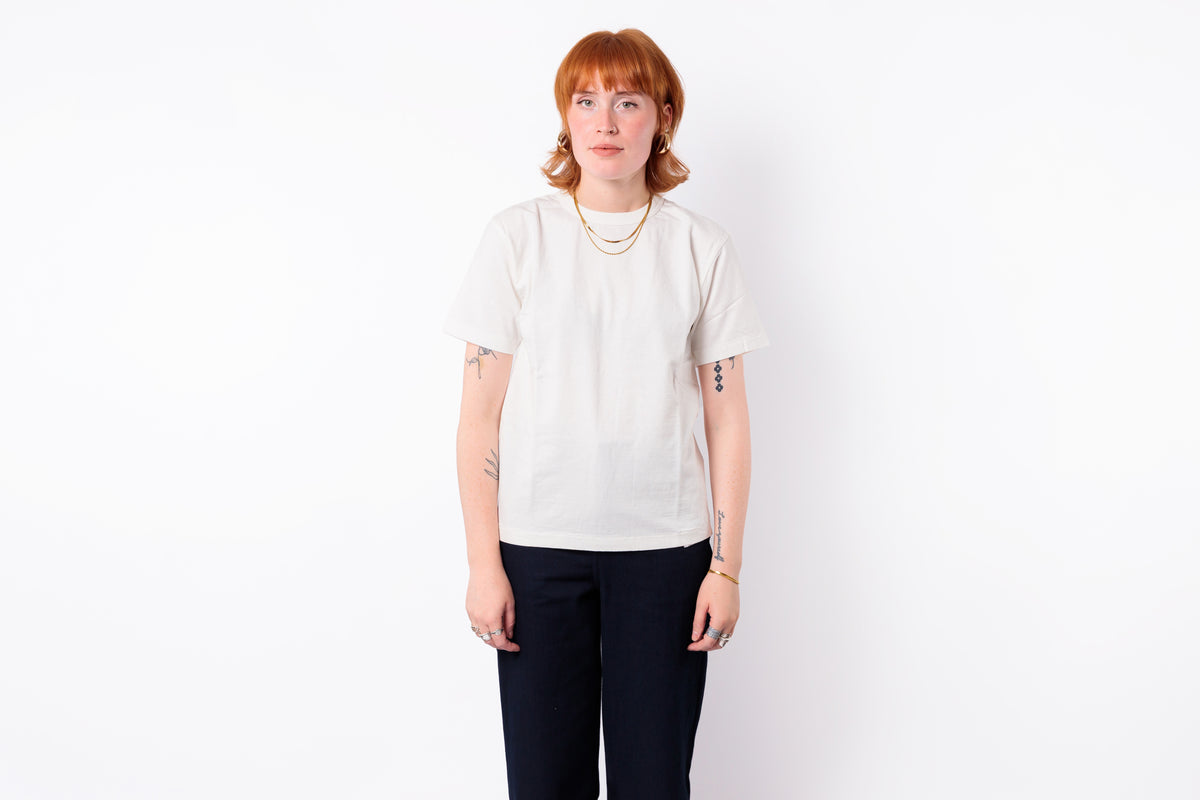 A model with short red hair stands against a plain white background wearing a cream-colored short-sleeve t-shirt and dark navy pants.
