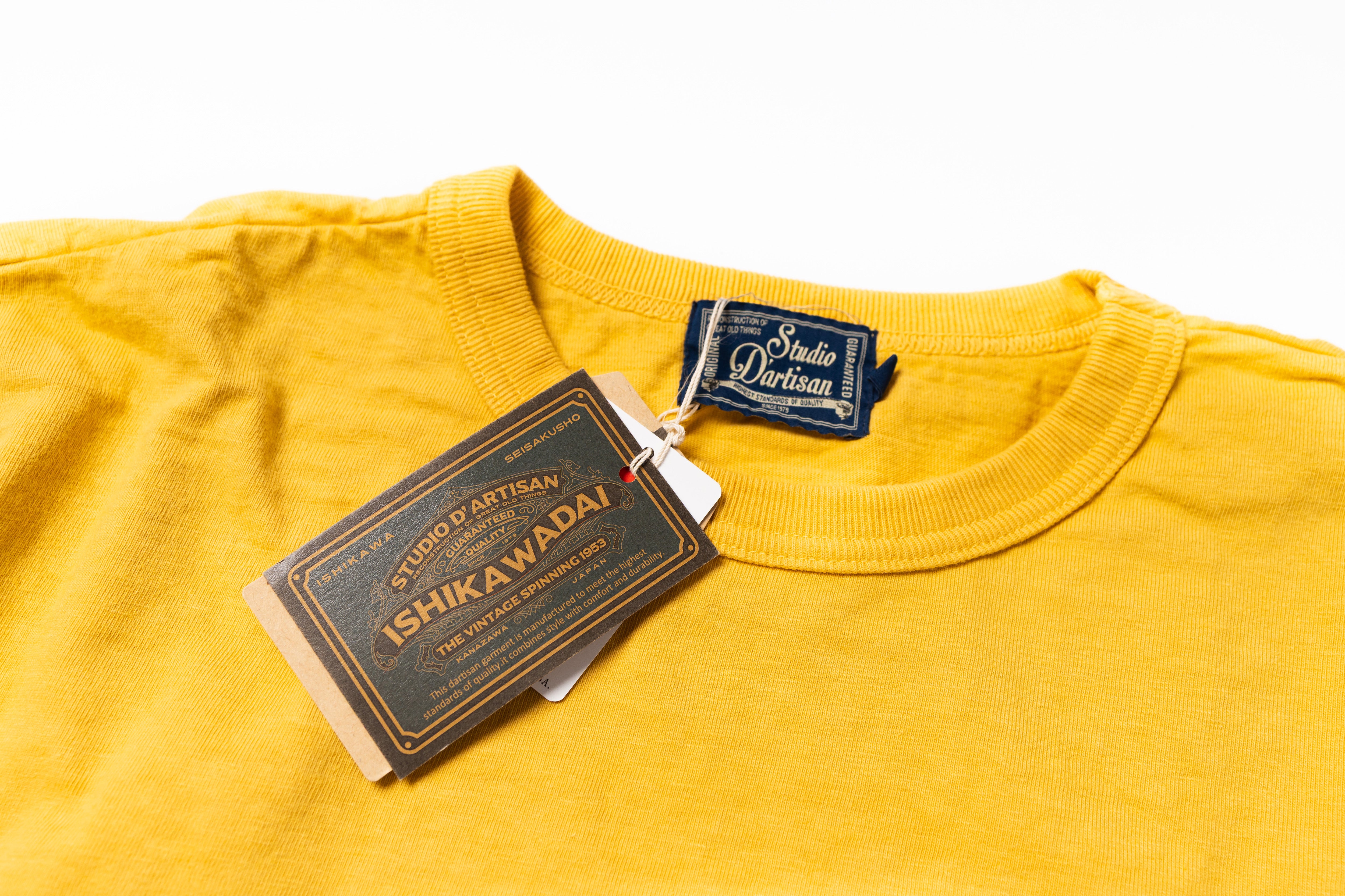 A close-up of a mustard yellow crewneck t-shirt by Studio D'Artisan, featuring a branded neck label and a hanging paper hangtag with 'Toshio D'Artisan' and 'Shikawadai' printed on it, set against a plain white background.