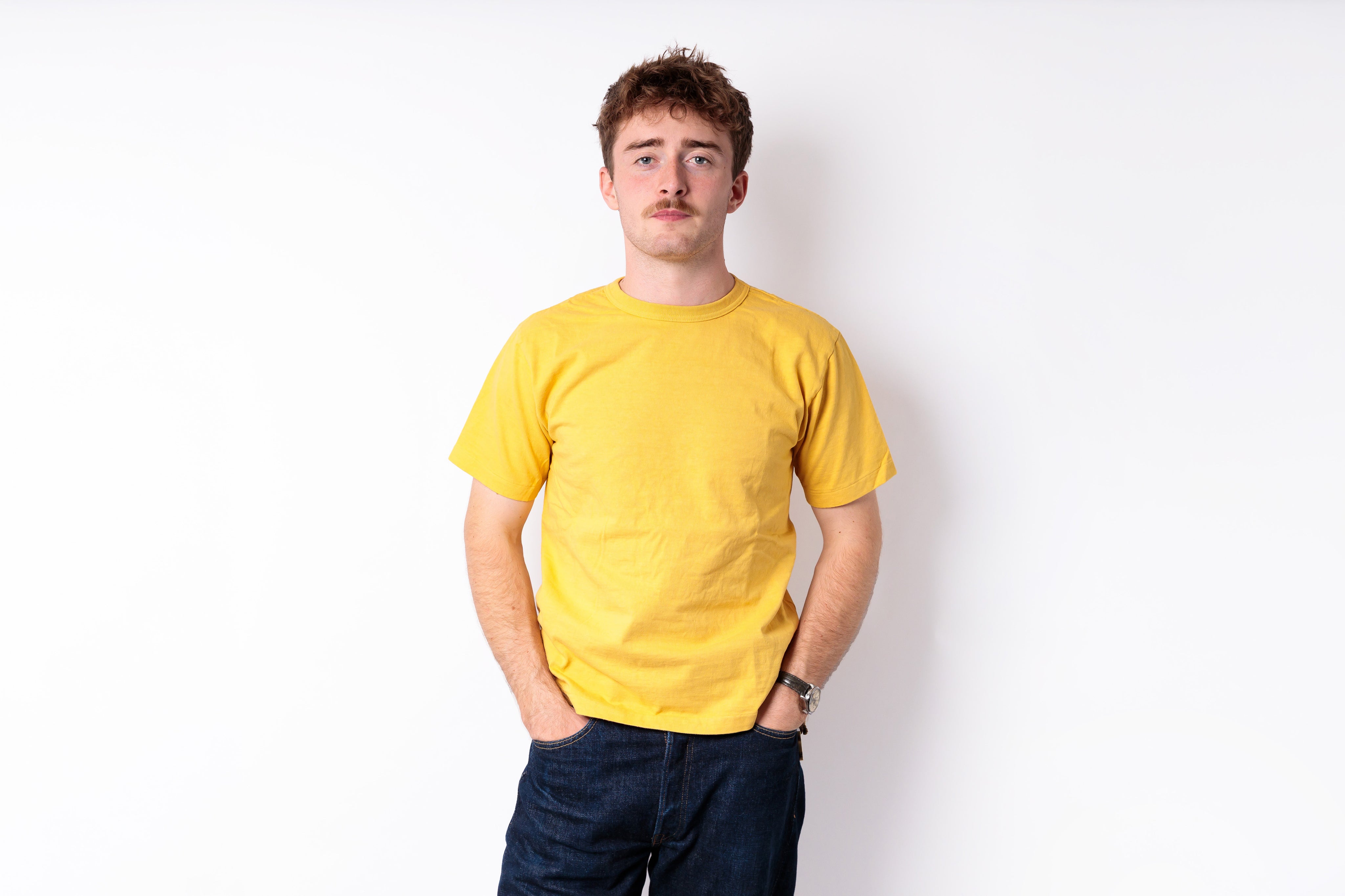 A man with a mustache wearing a plain yellow t-shirt and dark blue jeans, standing against a solid white background.