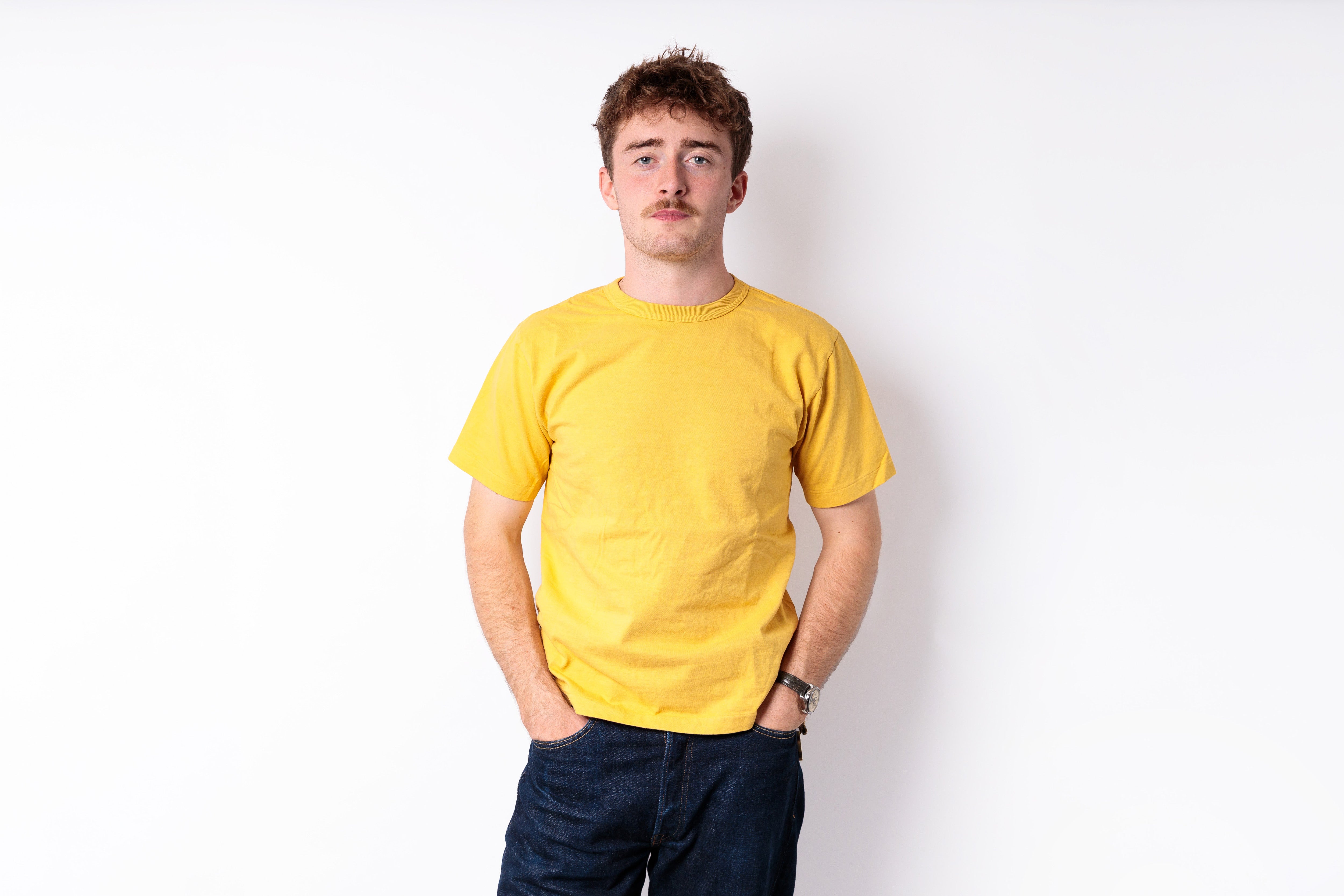 A man with a mustache wearing a plain yellow t-shirt and dark blue jeans, standing against a solid white background.