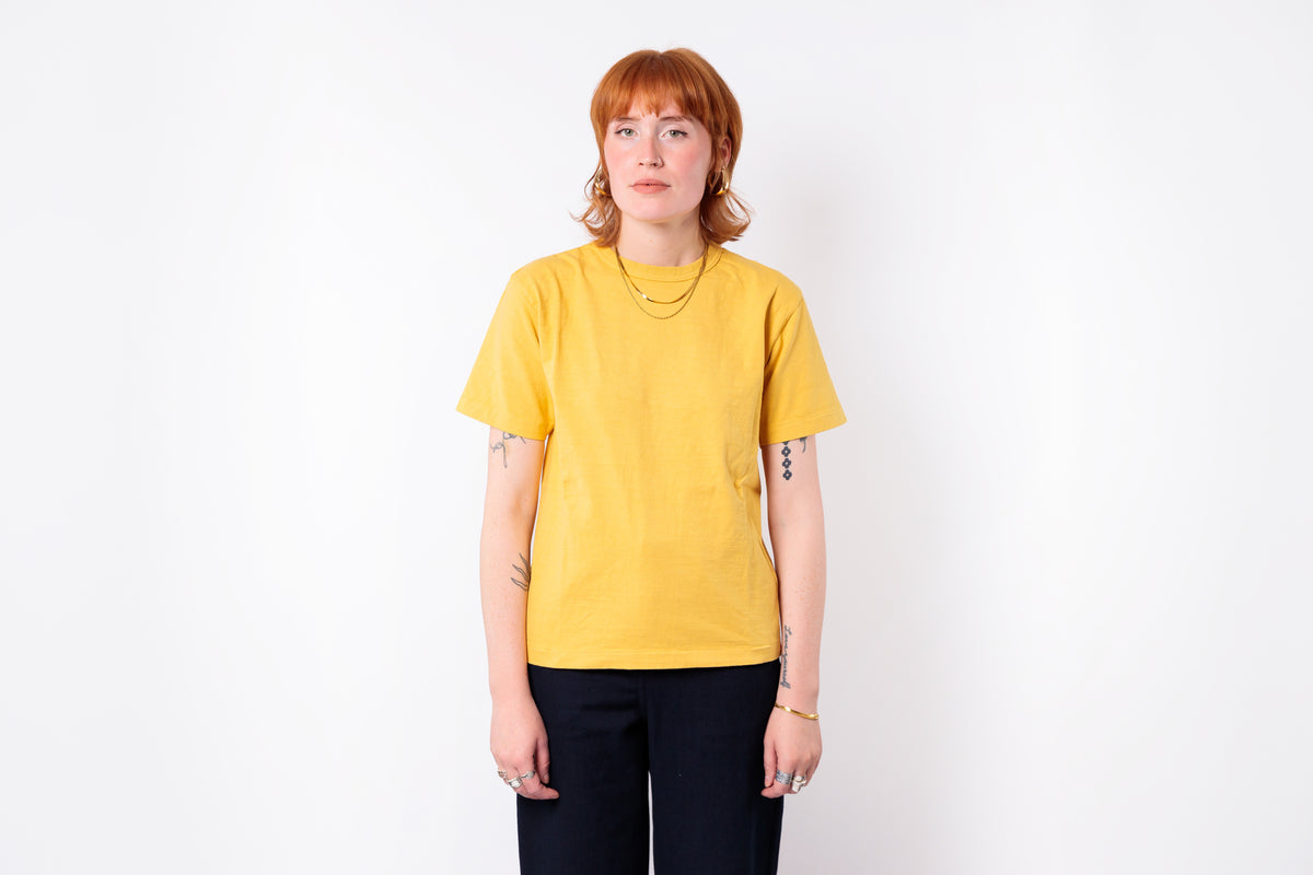 A woman with short red hair stands against a white studio background wearing a bright yellow short-sleeve crewneck t-shirt and dark navy pants.