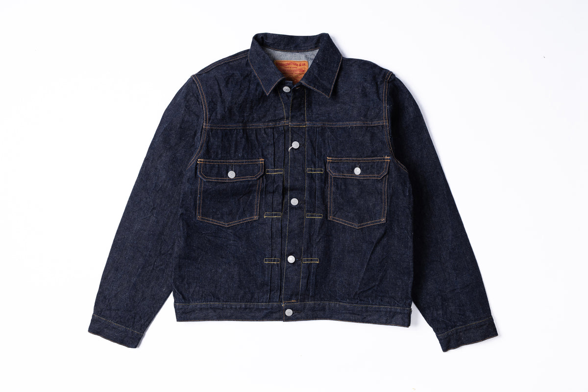 A dark indigo denim trucker jacket with contrast gold-toned stitching, silver snap buttons, and a classic collar, laid flat against a white background.