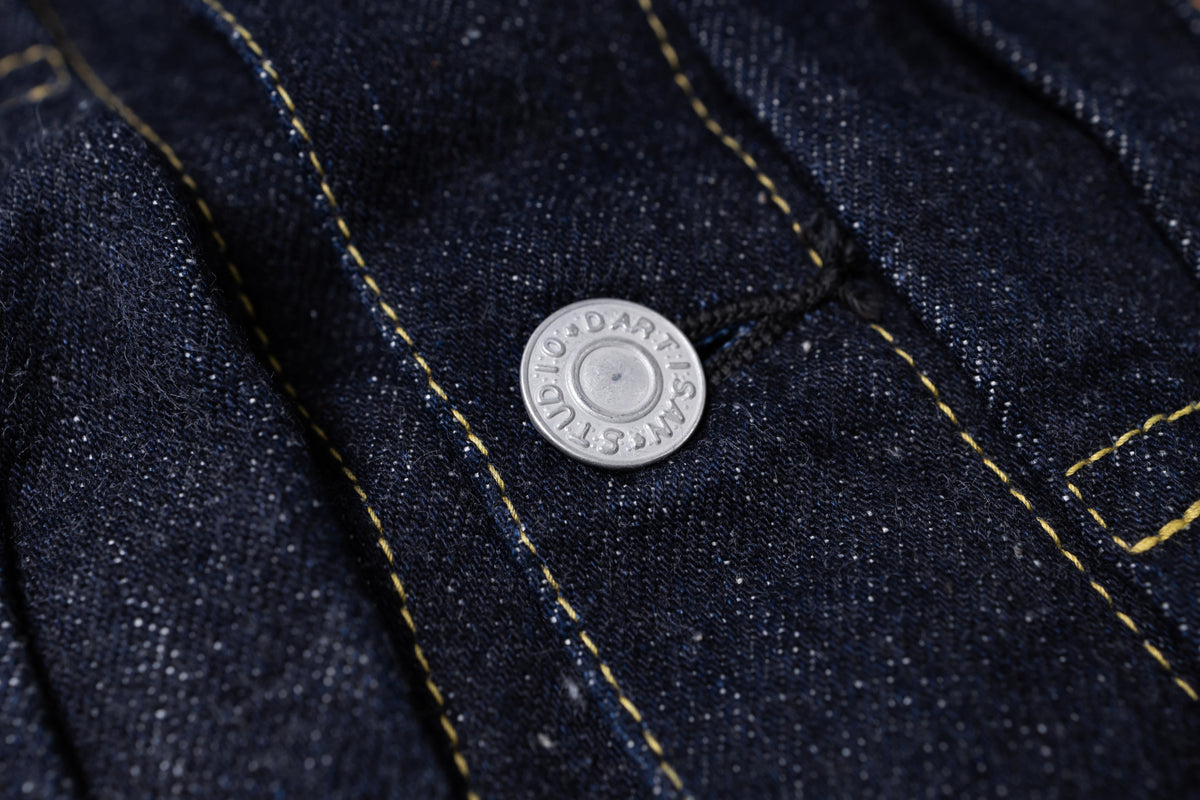 Close-up of dark indigo denim fabric with yellow contrast stitching and a round silver metal button embossed with the brand name.