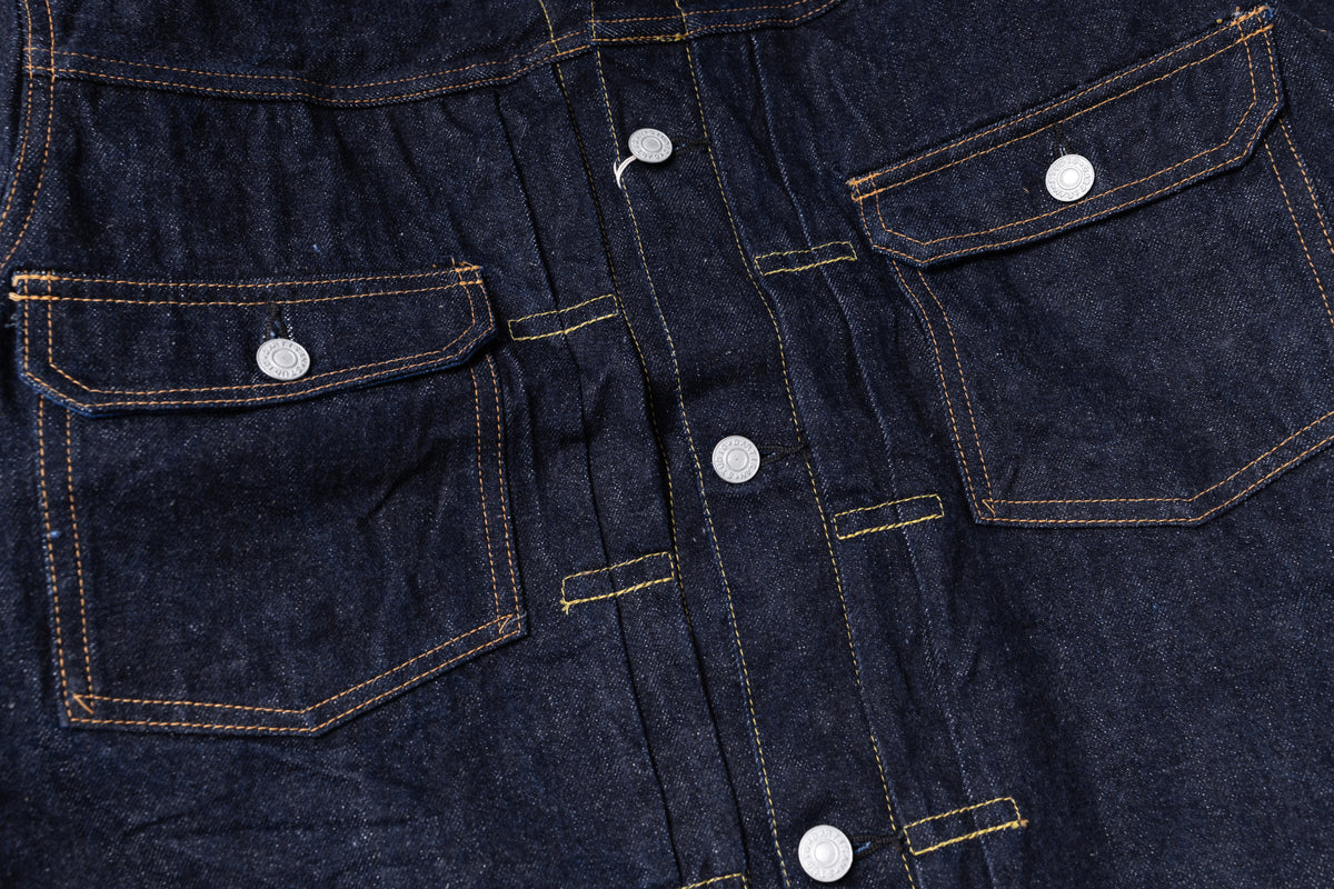 Close-up of dark indigo denim fabric with contrast tan stitching, silver metal buttons, and two flap chest pockets.