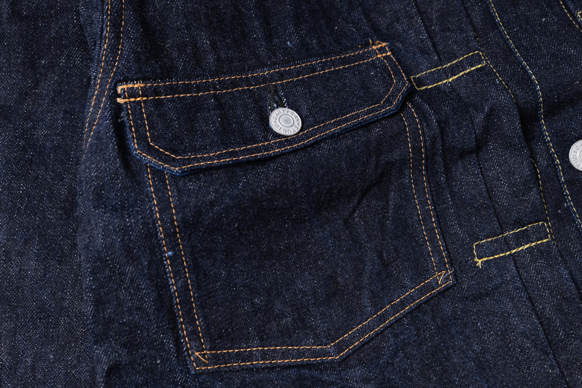 Close-up of a dark indigo denim garment showing a front flap pocket with contrast yellow-orange stitching and a silver metal button.