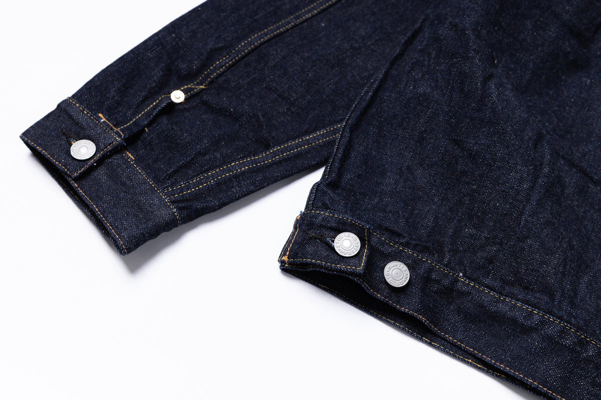 Close-up of a dark indigo denim jacket showing the buttoned cuff and hem detail with silver-toned buttons on a white background.