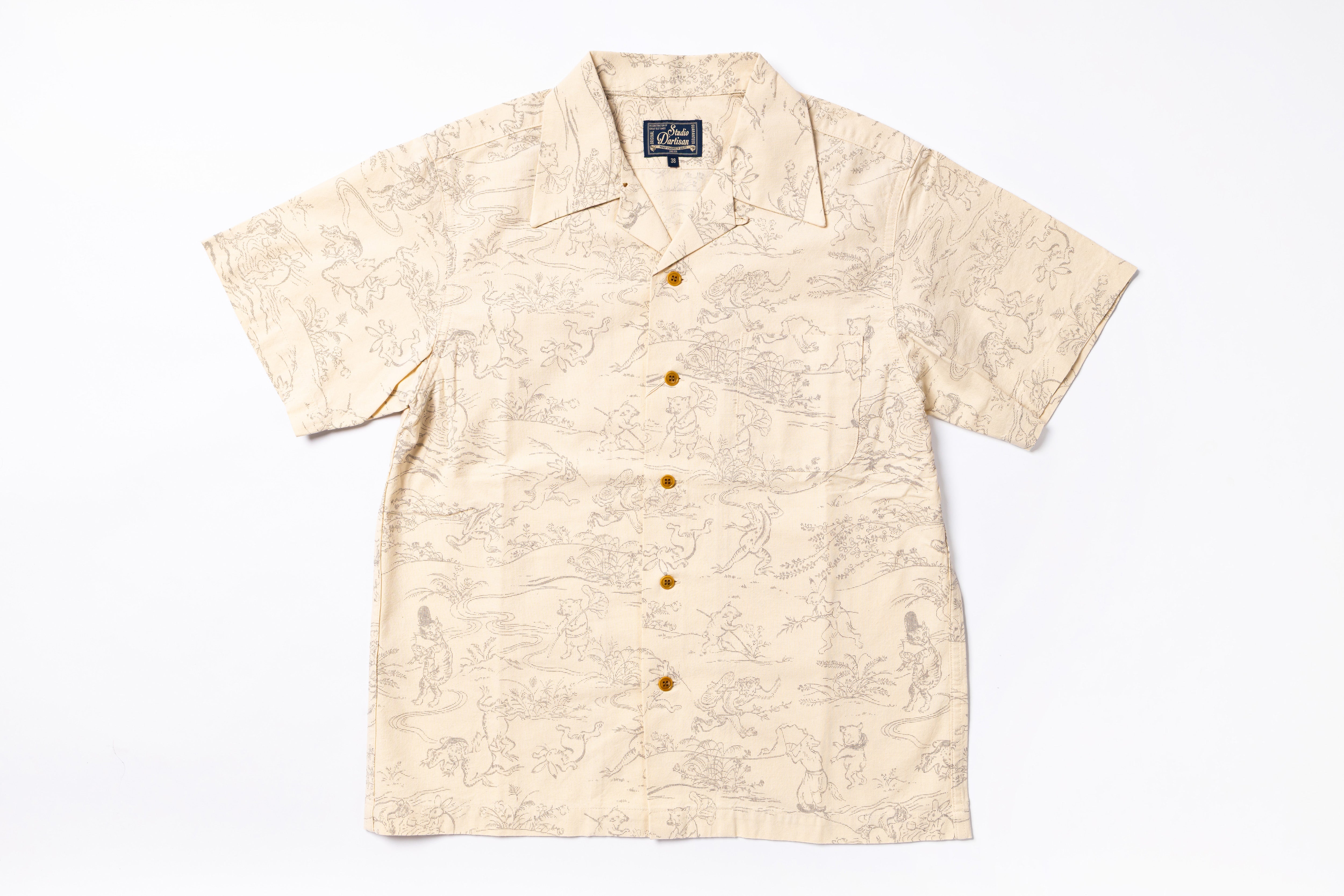 A short-sleeve camp collar button-up shirt in beige with a light brown scenic illustration pattern, featuring brown buttons down the front, laid flat on a white background.