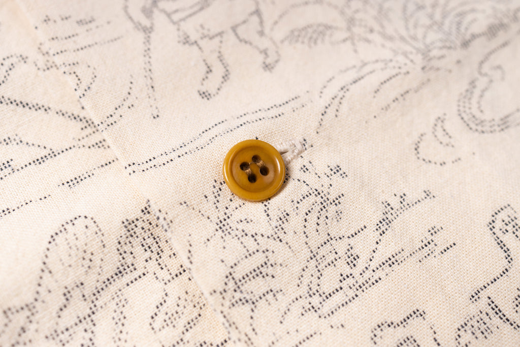 Close-up of a cream-colored fabric with a delicate black line-art pattern and a single round mustard-yellow button.