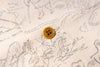 Close-up of a cream-colored fabric with a delicate black line-art pattern and a single round mustard-yellow button.