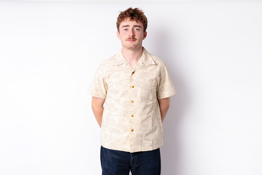 A man wearing a light beige short-sleeve button-up shirt with a subtle tropical pattern and dark blue jeans, standing against a plain white studio background.