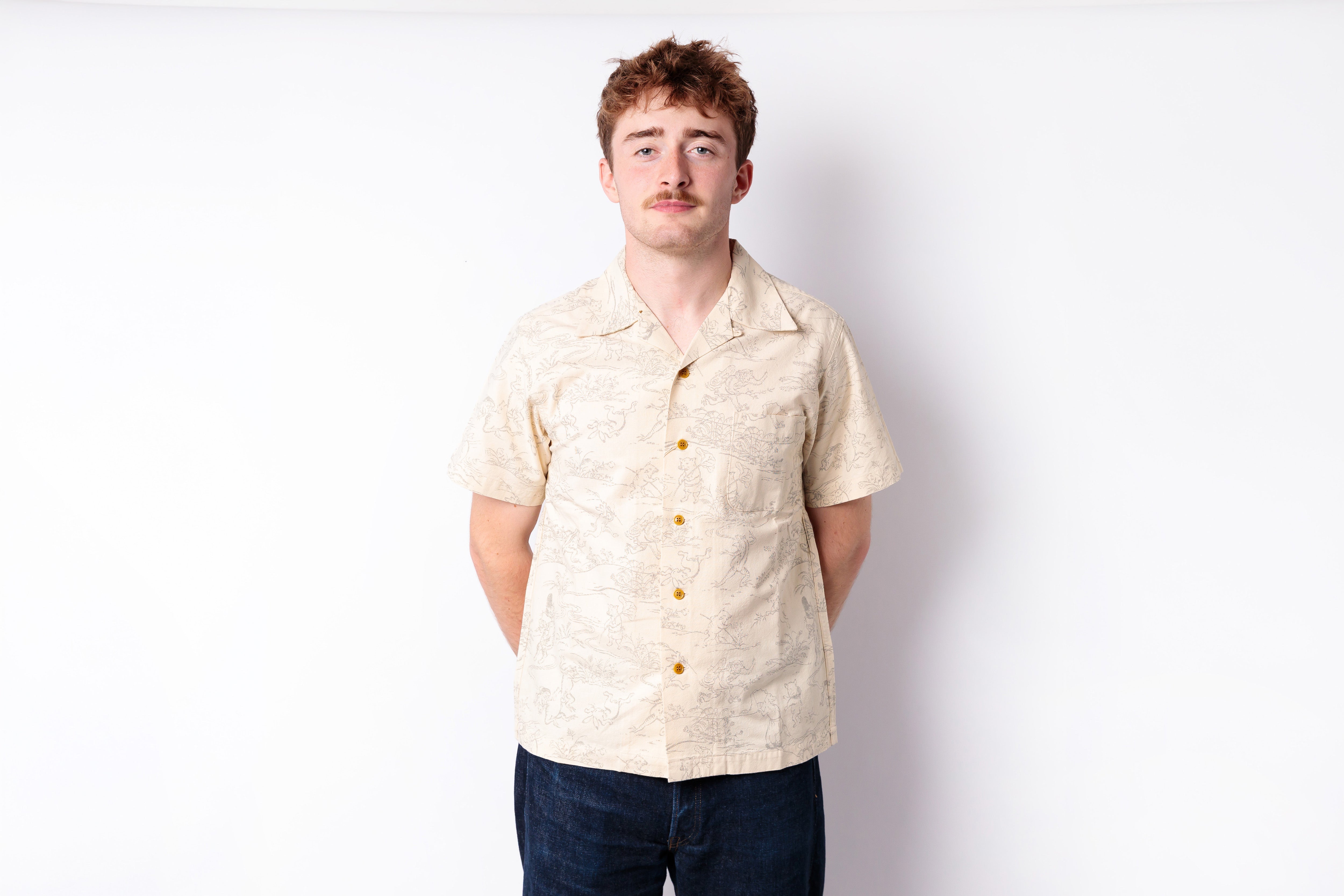 A man wearing a light beige short-sleeve button-up shirt with a subtle tropical pattern and dark blue jeans, standing against a plain white studio background.
