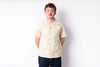 A man wearing a light beige short-sleeve button-up shirt with a subtle tropical pattern and dark blue jeans, standing against a plain white studio background.