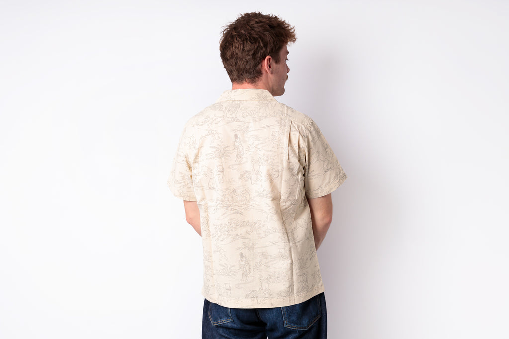 A man viewed from behind wearing a light beige short-sleeve button-up shirt with a subtle tonal pattern, paired with dark blue jeans, against a plain white background.
