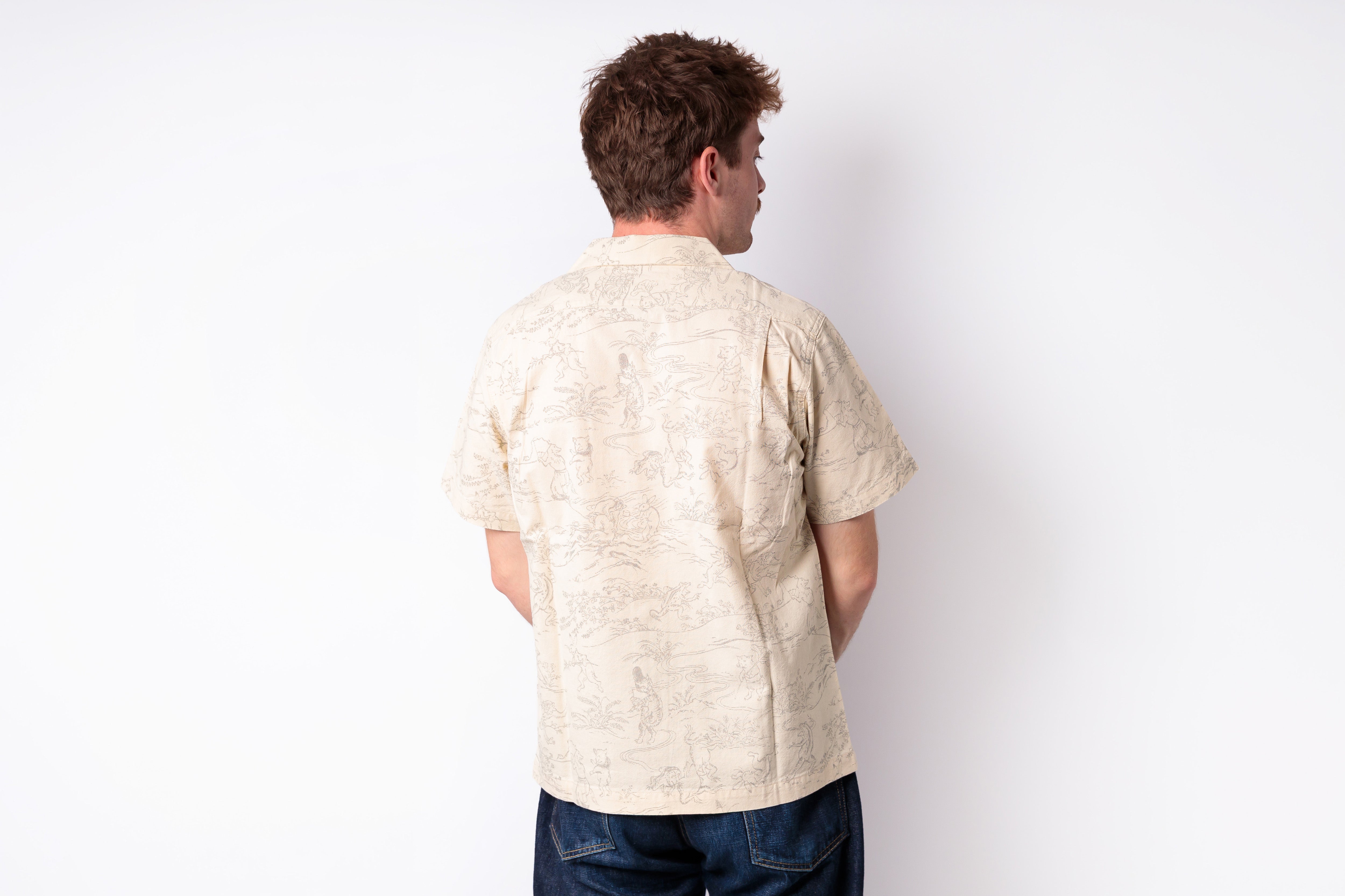 A man viewed from behind wearing a light beige short-sleeve button-up shirt with a subtle tonal pattern, paired with dark blue jeans, against a plain white background.