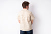 A man viewed from behind wearing a light beige short-sleeve button-up shirt with a subtle tonal pattern, paired with dark blue jeans, against a plain white background.