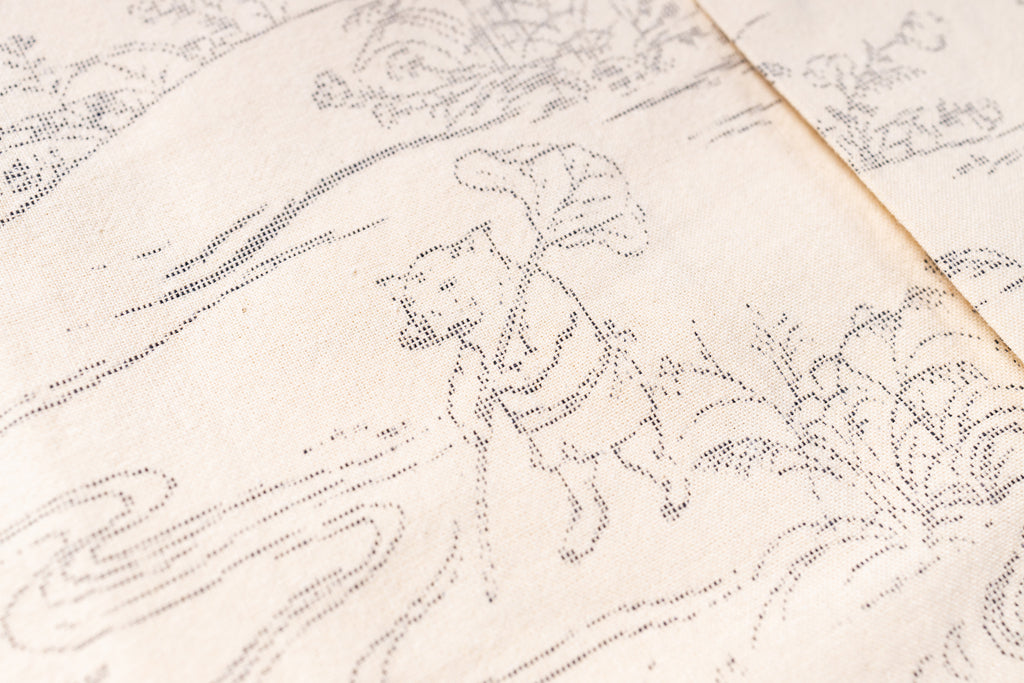 Close-up of a cream-colored fabric with a fine black dotted line illustration of a figure walking among stylized plants and waves.