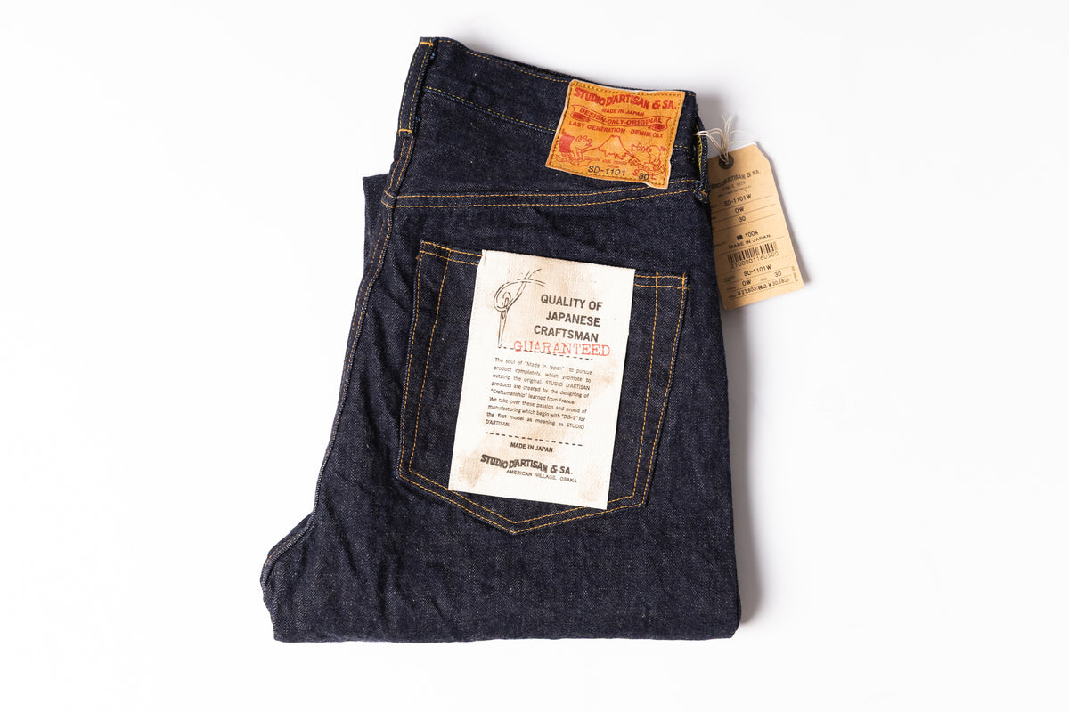 A folded pair of dark indigo denim jeans with a prominent white paper guarantee card on the back pocket, a small orange label, and a paper hangtag, displayed on a clean white background.