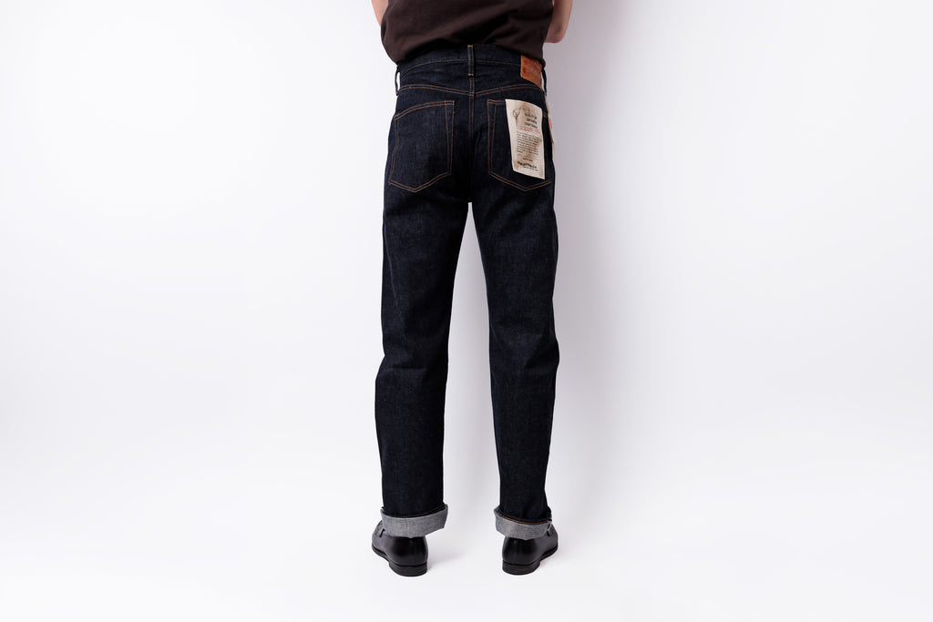 Rear view of a person wearing dark indigo denim jeans with cuffed hems and black shoes, standing against a plain white studio background.