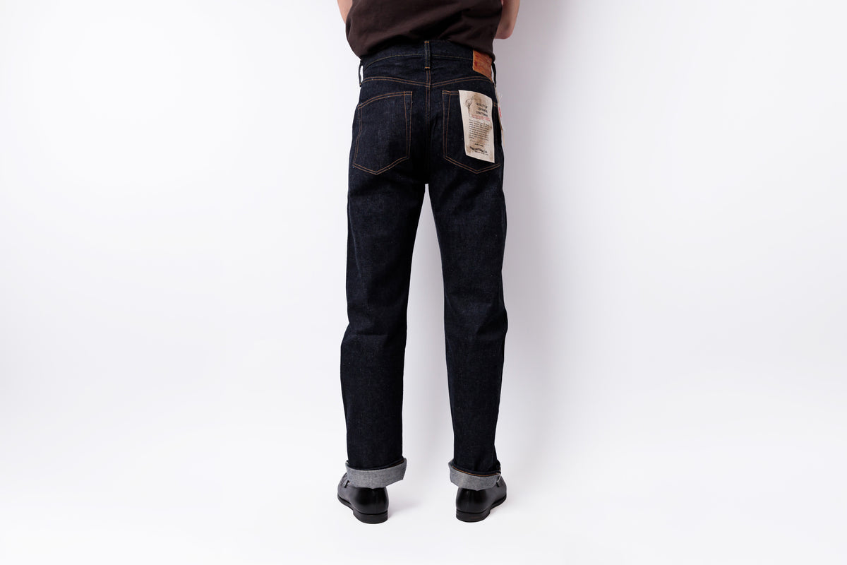Rear view of a person wearing dark indigo denim jeans with cuffed hems and black shoes, standing against a plain white studio background.