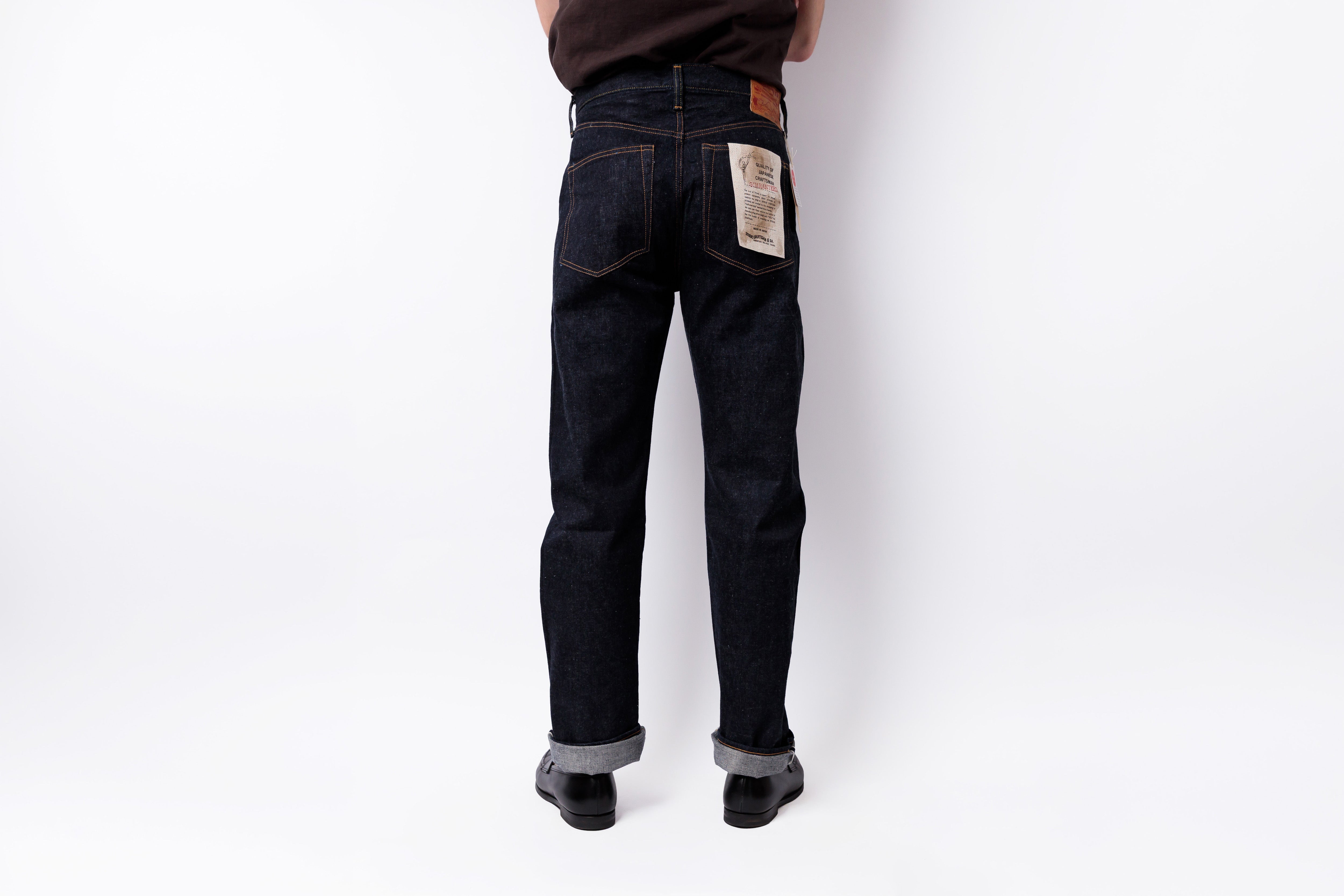 Rear view of a person wearing dark indigo denim jeans with cuffed hems and black shoes, standing against a plain white studio background.
