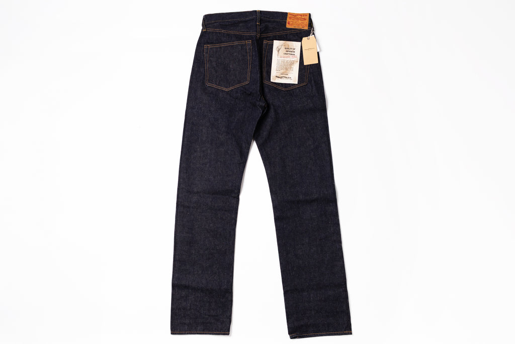 Dark indigo denim jeans shown from the back, featuring a classic five-pocket design, a leather patch at the back waistband, and paper tags attached to the rear pockets, set against a plain white background.