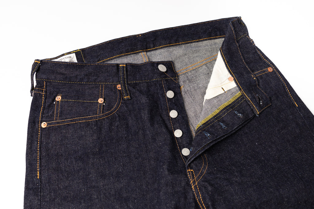Close-up of a pair of dark indigo denim jeans with a button fly and contrast orange stitching, shown on a white background.