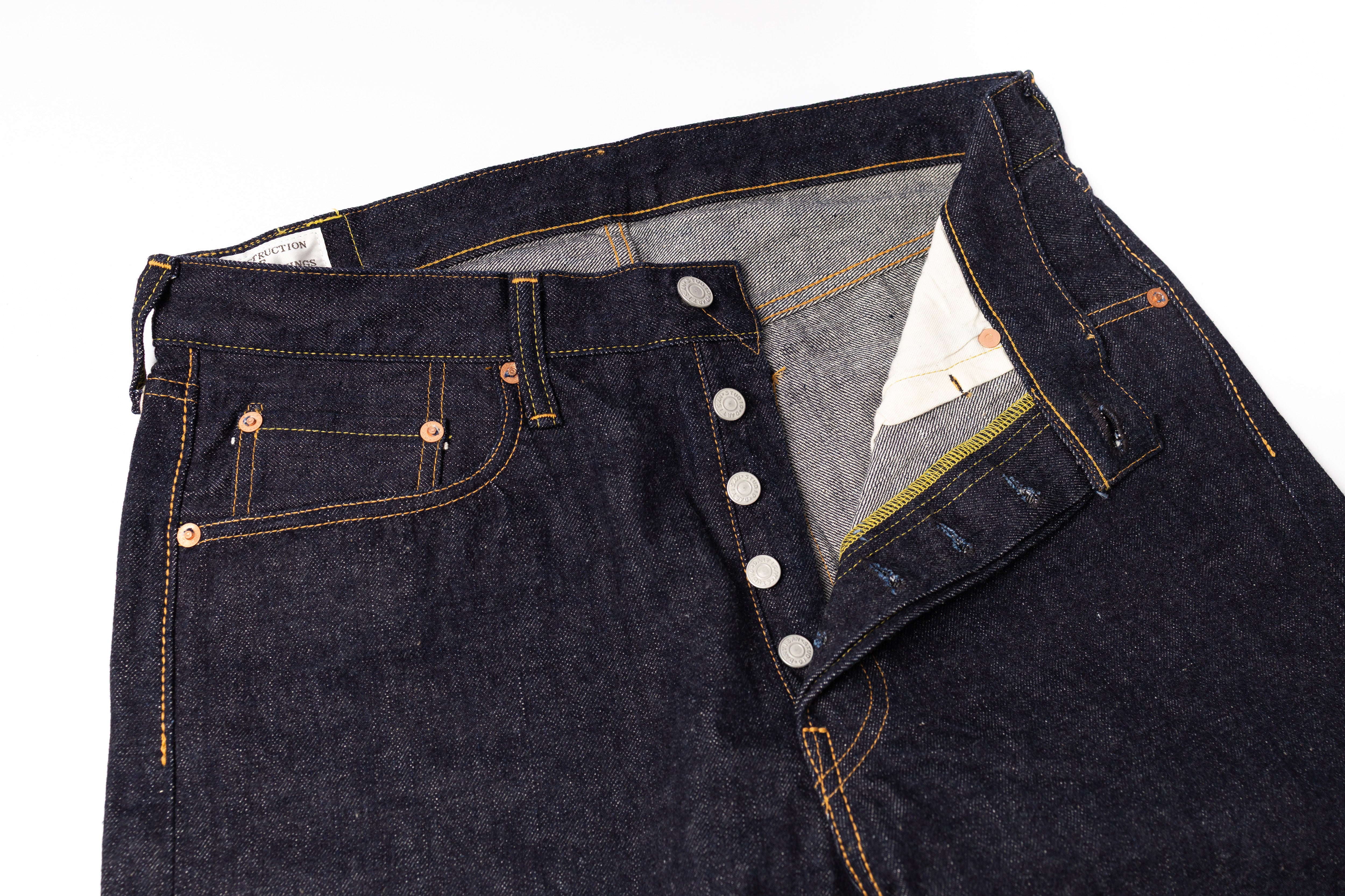 Close-up of a pair of dark indigo denim jeans with a button fly and contrast orange stitching, shown on a white background.