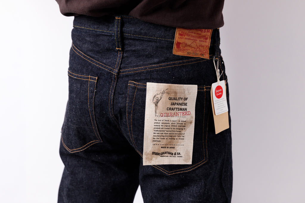 Close-up back view of a person wearing dark indigo denim jeans with a tan leather patch and a paper hangtag attached to the back pocket, against a plain light background.