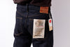 Close-up back view of a person wearing dark indigo denim jeans with a tan leather patch and a paper hangtag attached to the back pocket, against a plain light background.