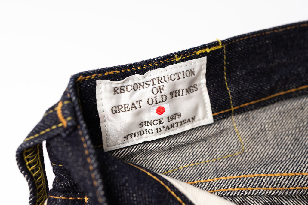 Close-up of the inner waistband of dark indigo denim jeans, showing a white woven label that reads 'RECONSTRUCTION OF GREAT OLD THINGS', 'SINCE 1979', and 'STUDIO D'ARTISAN' with a small red dot, alongside yellow contrast stitching.
