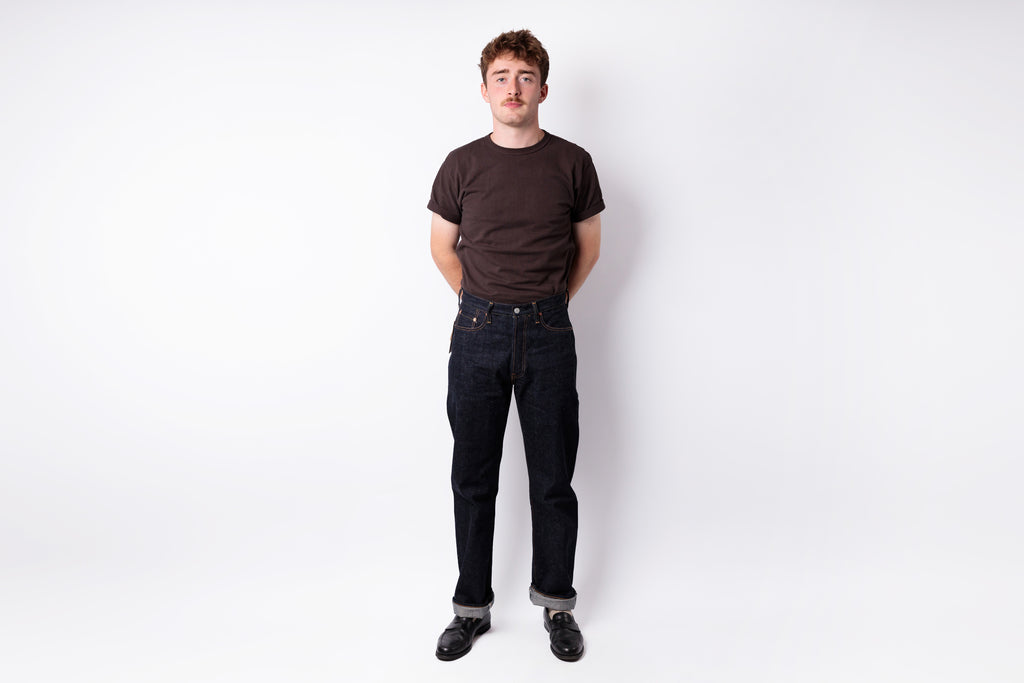 A young man standing against a white studio background, wearing a dark brown crewneck t-shirt, dark blue jeans, and black shoes.